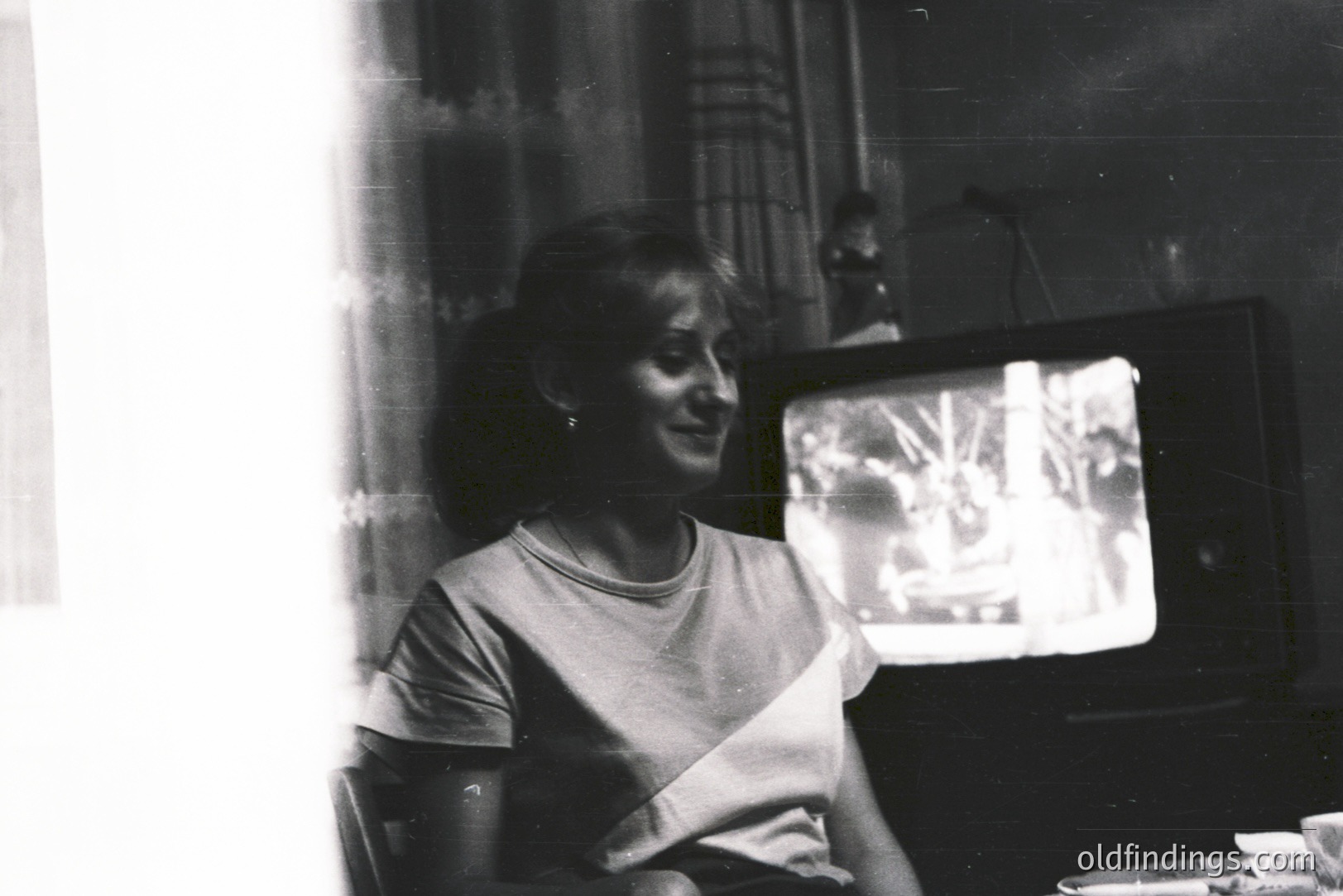 Woman seated, watching television in a dimly lit room. 1970s style furniture and a large, wood-grain television suggest the era. Grainy black and white film; likely a candid snapshot. A figure is visible in the background.