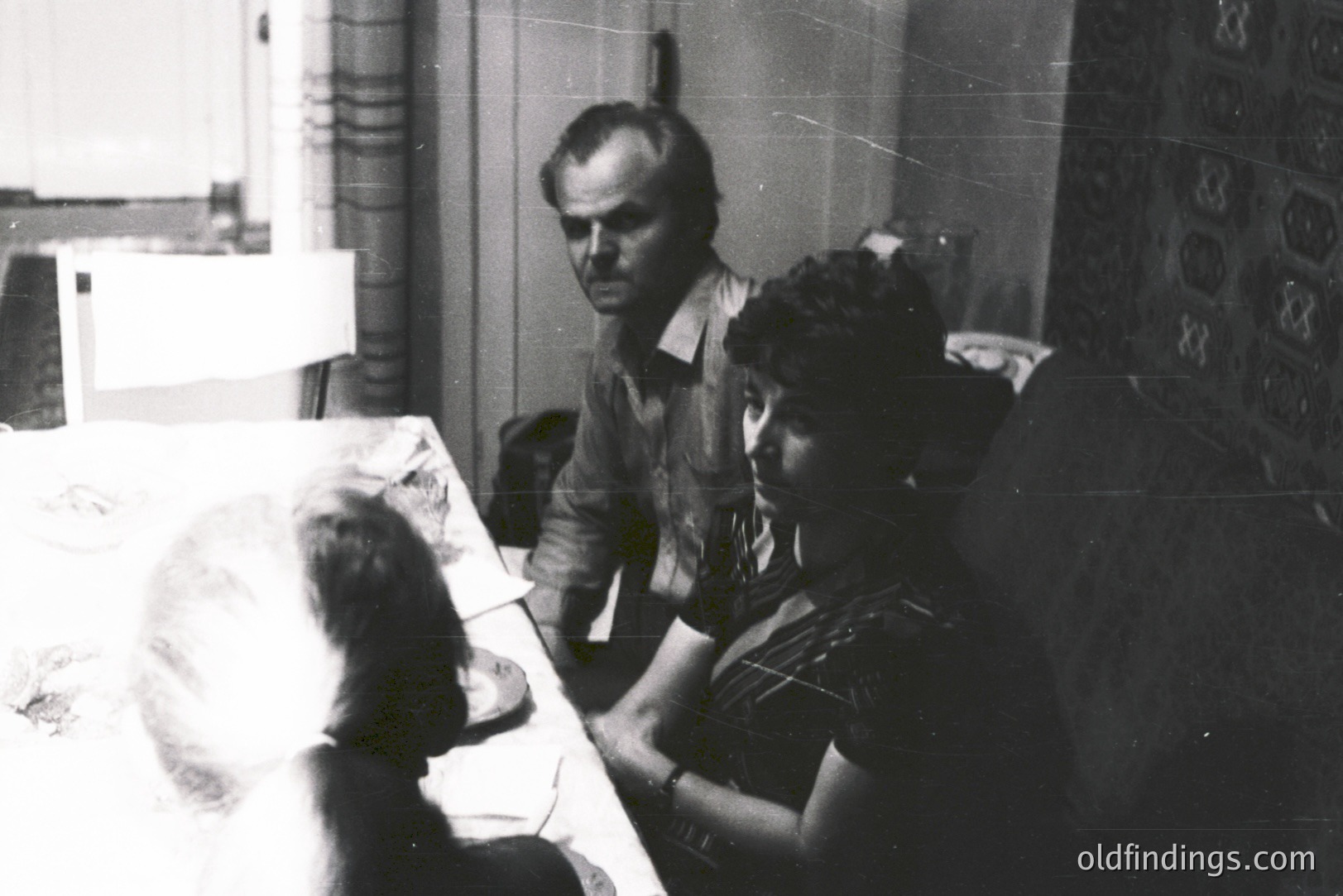A man in a plaid shirt observes a woman seated at a table covered with a patterned cloth. A partial view of a young child is seen at the table's edge. Interior scene with visible wallpaper and a window. Likely a domestic setting, circa 1970s. A candid, personal moment.