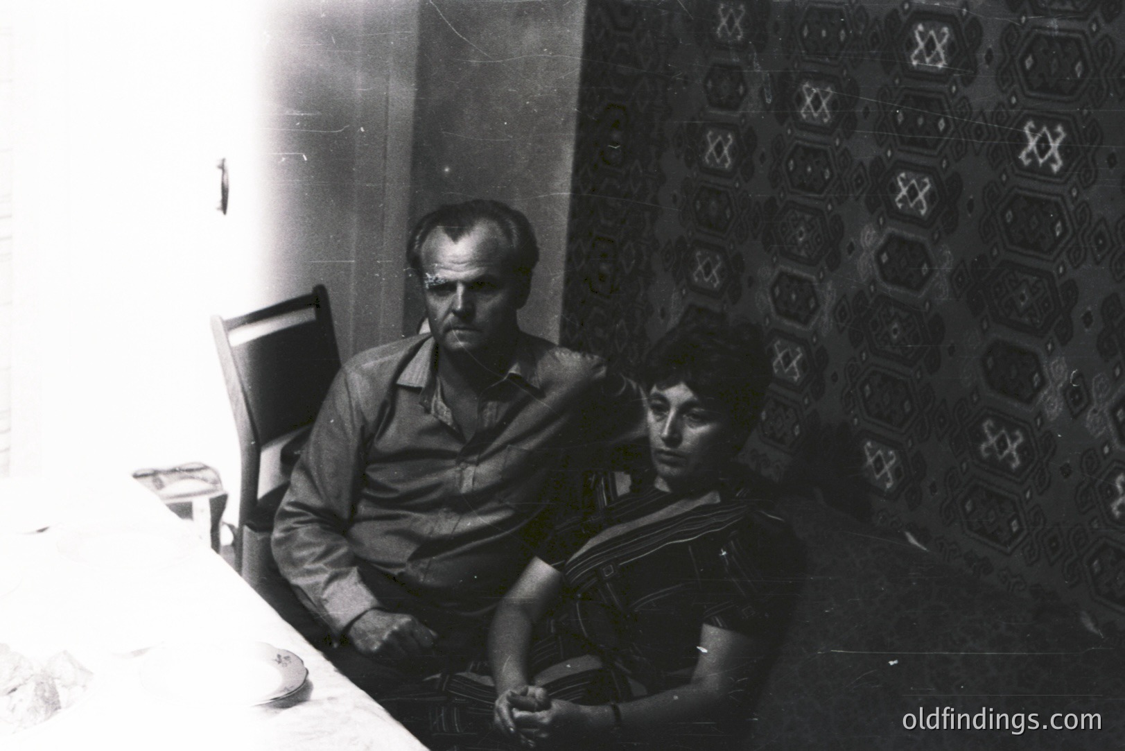 A black and white portrait shows two men seated in a courtyard, likely in Eastern Europe. The man on the left exhibits weathered features and a stern expression. The other man has short, dark hair and appears to be looking downwards. Decorative tilework adorns the wall behind them. Likely 1970s, indicative of socialist-era architecture.