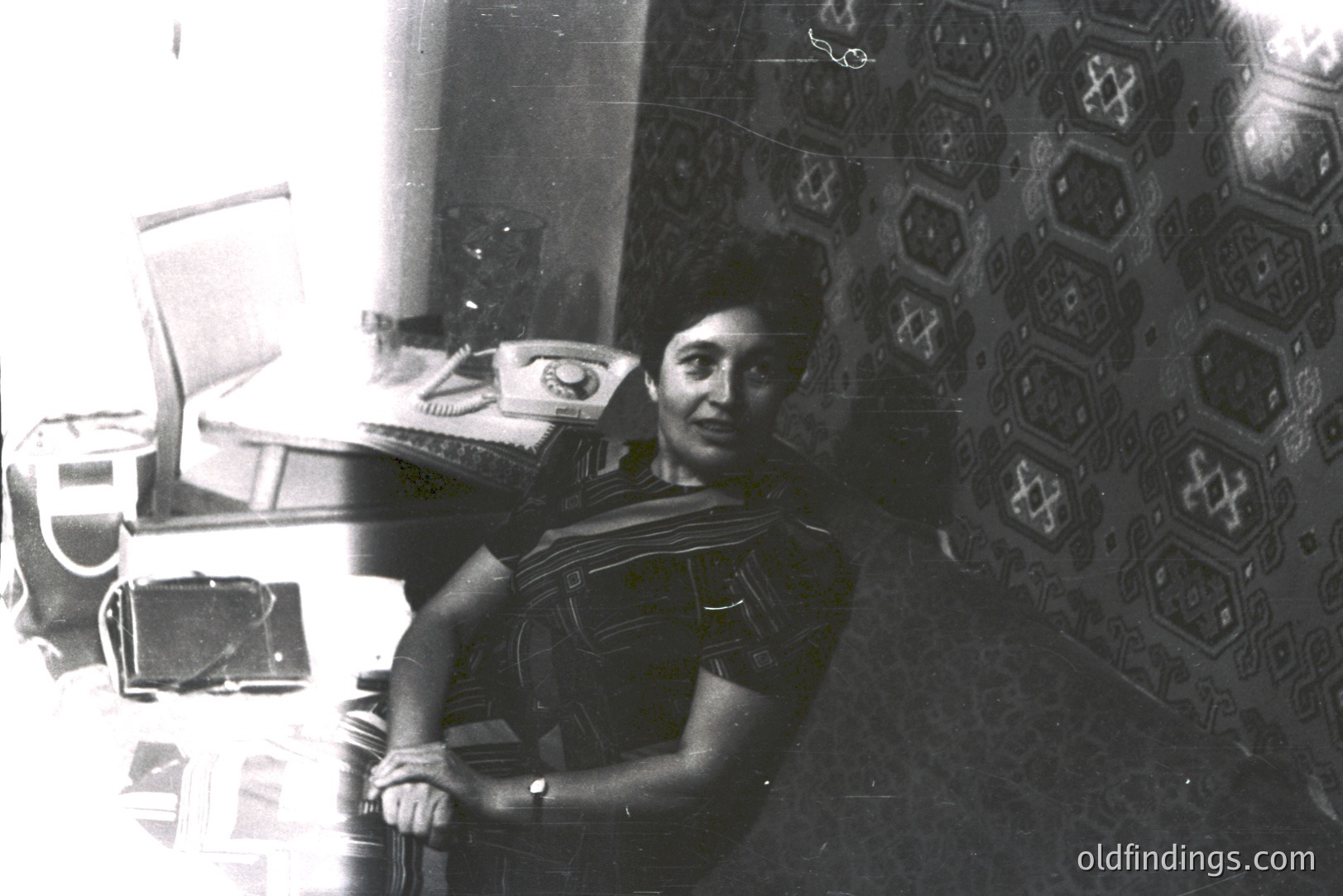 A seated woman in a patterned, sleeveless garment smiles directly at the camera. She is positioned within a room with patterned wallpaper and what appears to be a vintage telephone on a nearby table. Appears to be a candid portrait. Likely 1960s-1970s.
