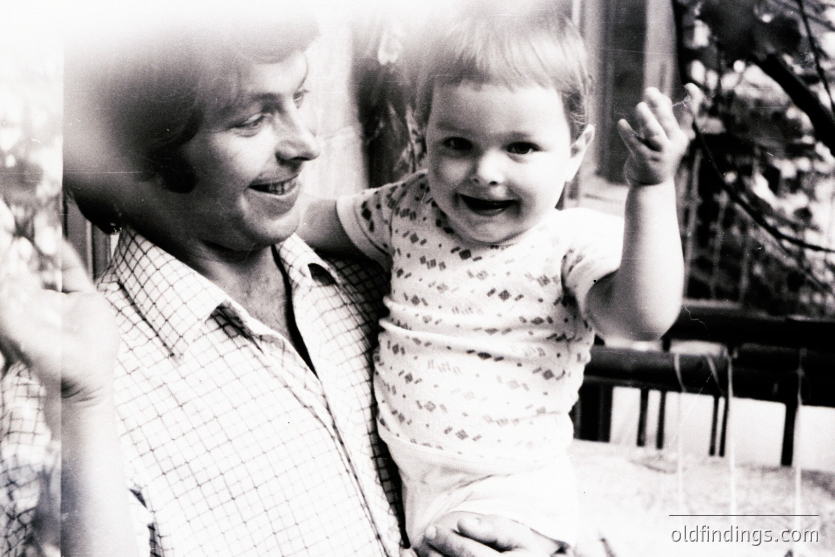 A smiling man holds a baby aloft, both gazing towards the camera. The man wears a plaid shirt; the baby, a patterned onesie. Likely taken outdoors, foliage is visible in the blurred background. A joyful, candid moment capturing familial connection. Appears to be a snapshot from the 1970s.