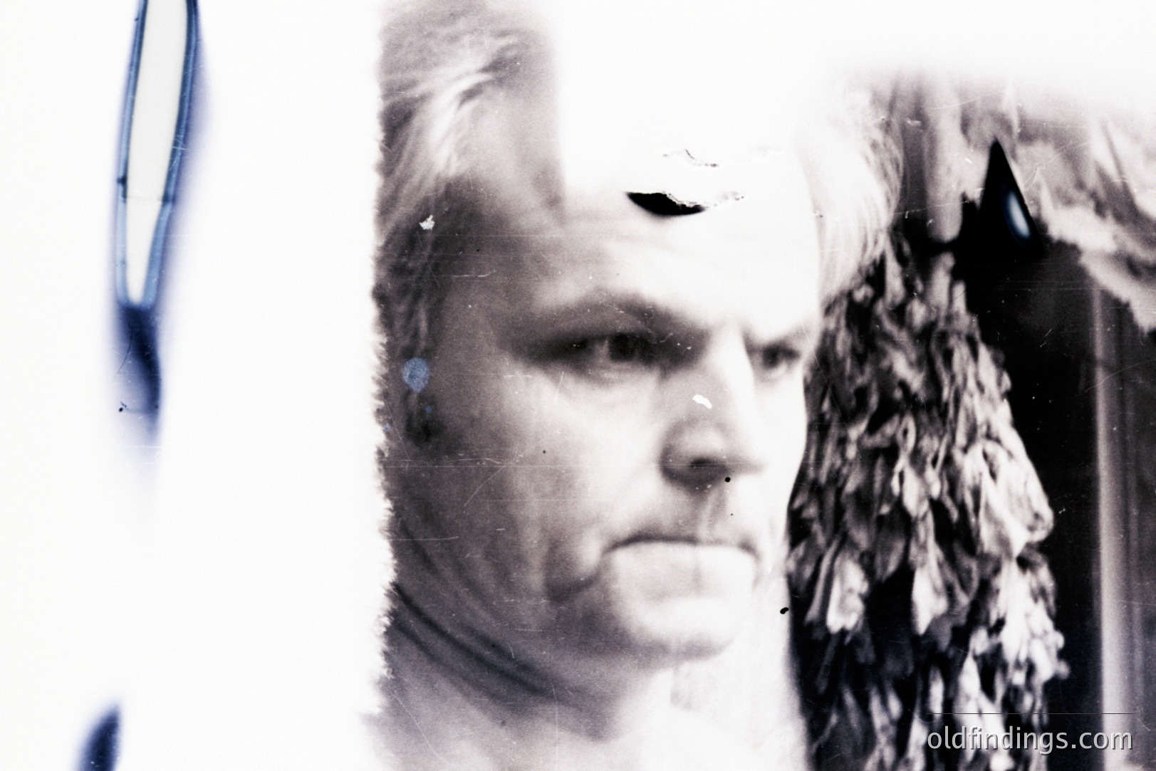 A close-up, black and white portrait of a man with short, styled hair and prominent facial features. The image shows signs of age and wear, possibly a degraded or damaged print. A blurred, textural element resembling foliage or dense branches is visible to the right. Appears to be a vintage studio portrait, potentially 1970s or 80s.