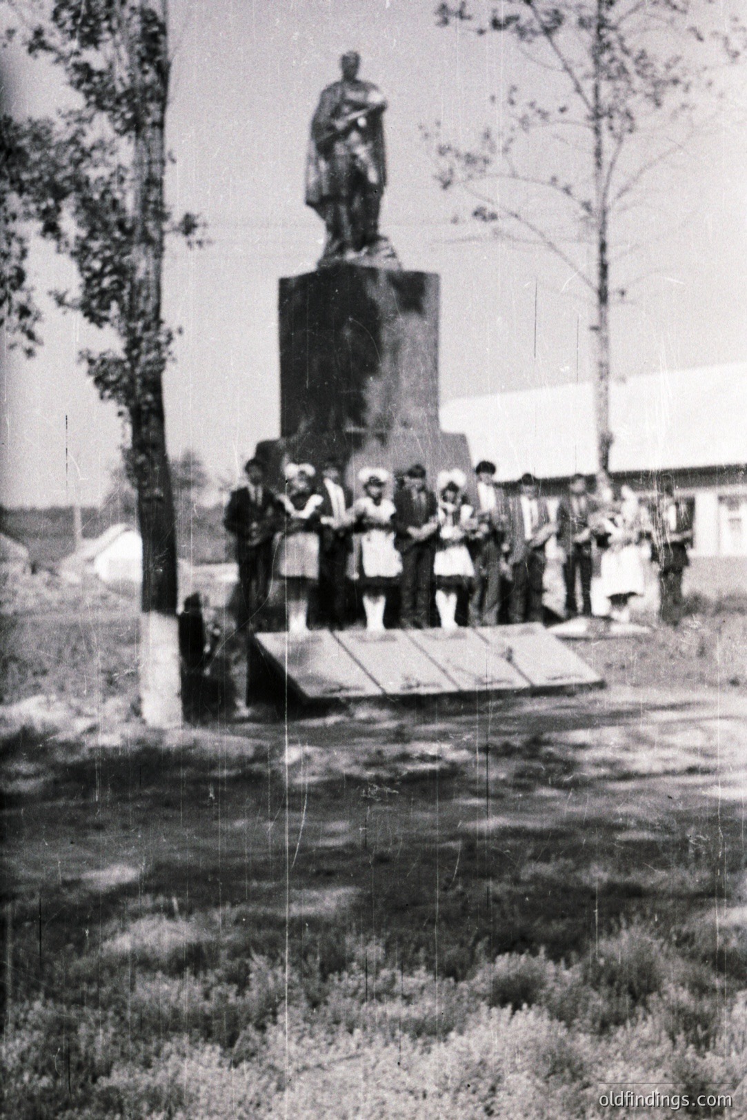 Monument stands in a village square with a group of formally dressed children and adults gathered at its base. The statue depicts a figure in military attire. Buildings and foliage form a backdrop, suggesting a rural setting. Likely Bulgaria, mid-20th century.