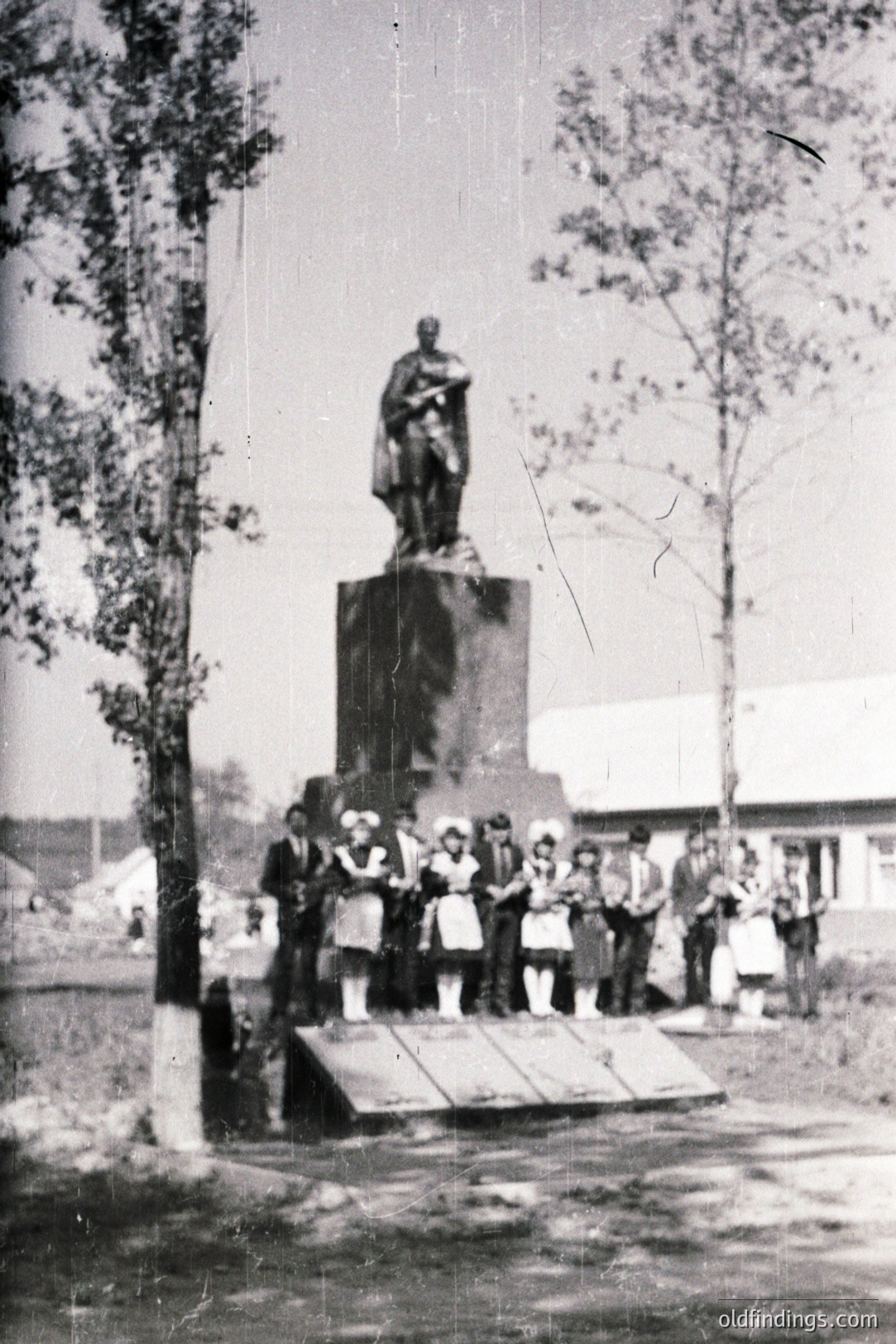 Monumental bronze statue atop a stone pedestal, viewed from a low angle. A group of formally-dressed children and adults stand on a raised platform in front. Likely a commemorative or national figure. Architectural background suggests a rural or small-town setting. Appears to be 1950s-1970s Eastern Europe.