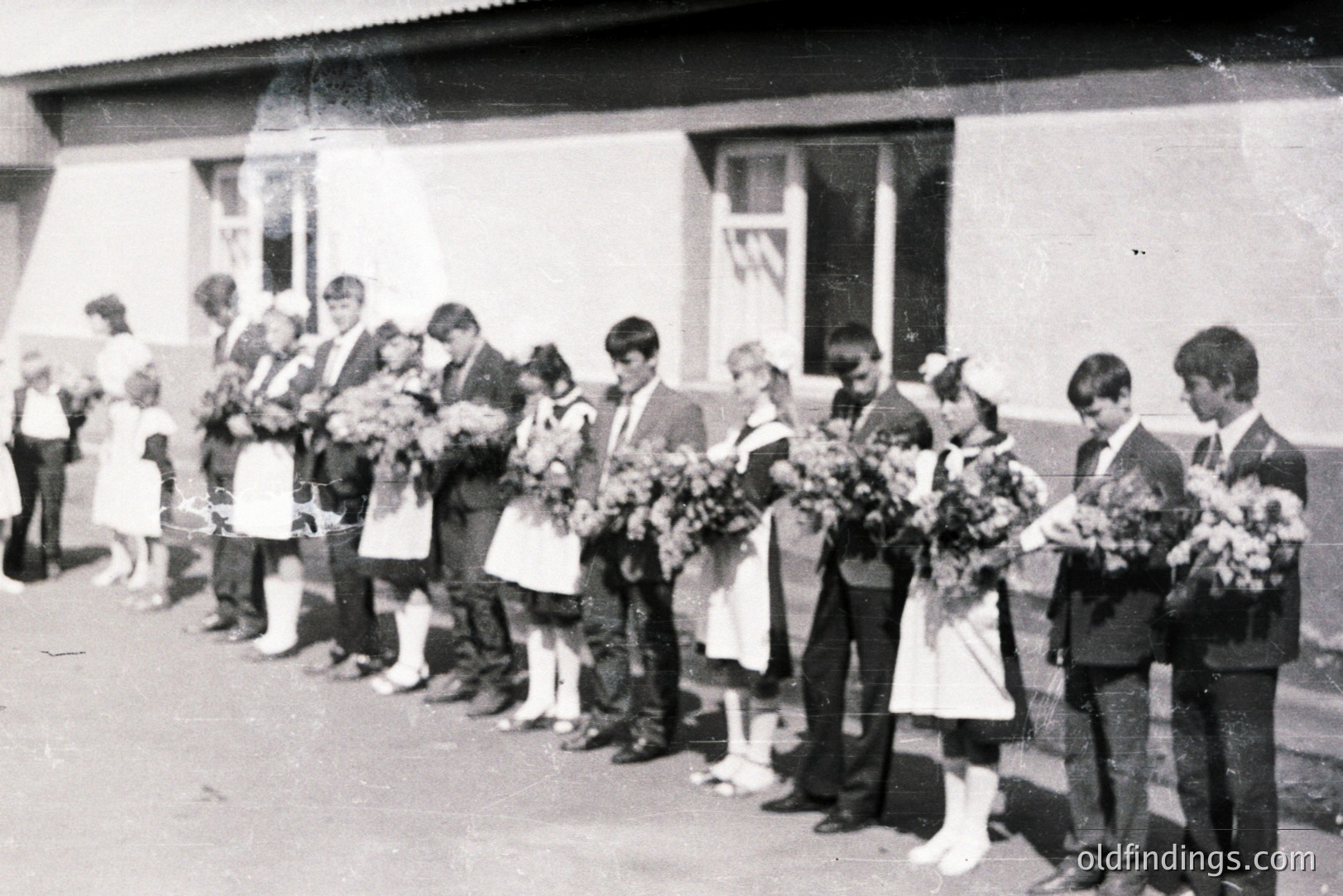 Group portrait of students, likely a graduating class. Boys wear dark suits & ties, girls wear white dresses with aprons. Each holds a bouquet of flowers. Building with plain facade visible behind. Appears to be 1960s, Eastern European setting. Valuable for historical research.