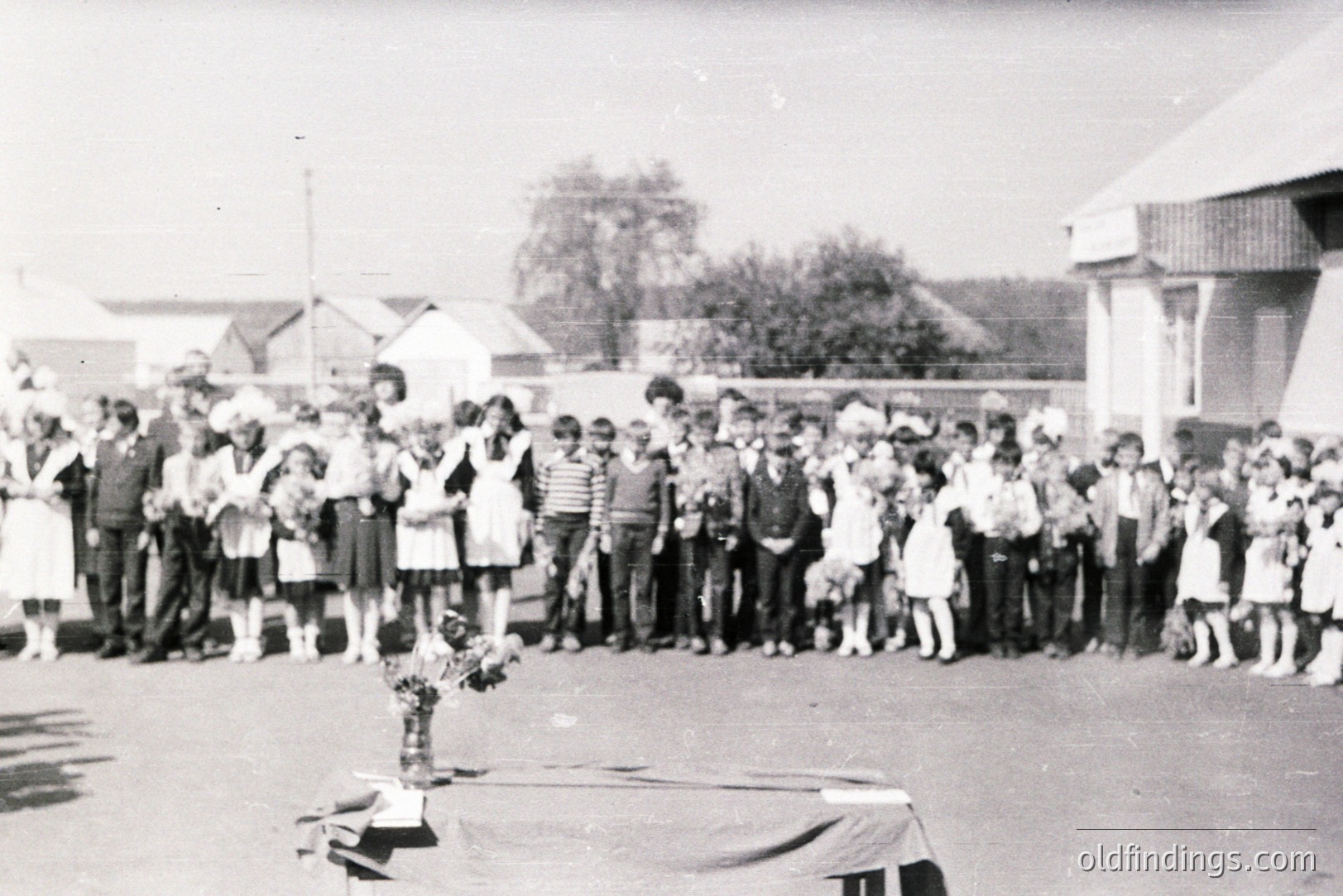 Group portrait of formally-dressed children, possibly a school class, gathered in a courtyard with residential buildings in the background. Likely Eastern Europe, 1960s-1970s. A simple table with floral arrangement is visible in the foreground. Demonstrates social structures & childhood.