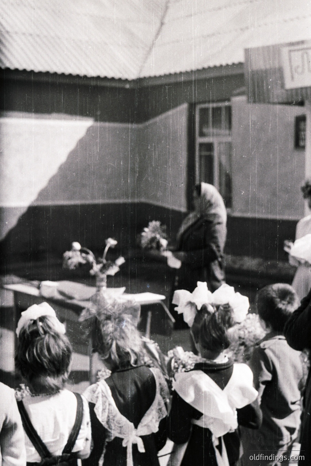 Group of children, likely school attendees, wear floral headwear and aprons in an outdoor courtyard setting. An adult figure stands nearby. Architectural style suggests Central/Eastern Europe. Appears to be a commemorative or celebratory event, possibly mid-20th century (1950s-1970s). Limited detail hinders definitive location ID.