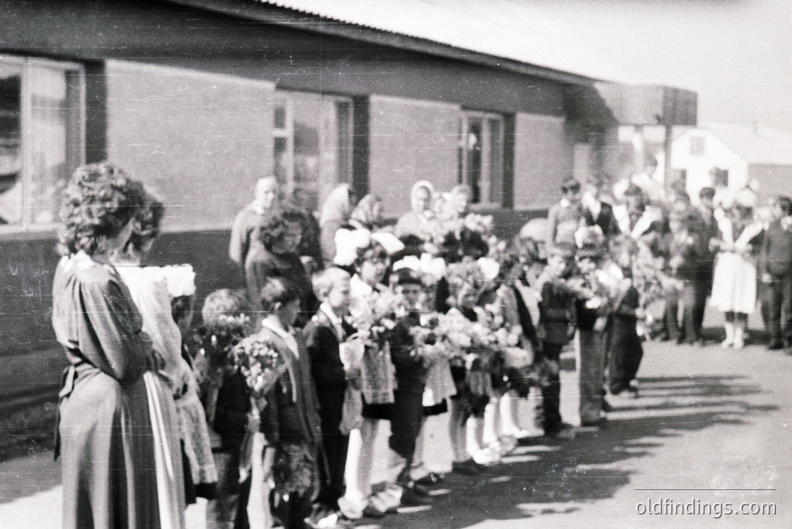 A line of schoolchildren, many holding flowers, stands before a low-slung building with large windows. A woman in a long dress observes them. Likely a school event or ceremony; details suggest a rural or small-town setting. Appears to be from the mid-20th century (1950s-1960s). A moment of community and childhood.