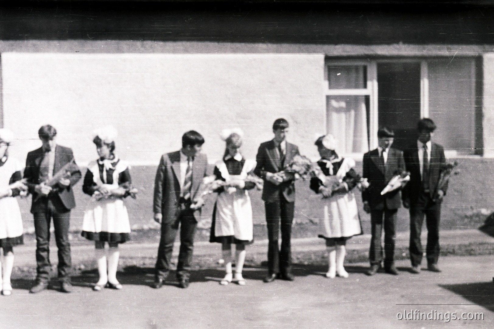 A group of six young people pose outside a simple, modernist building. Four wear traditional folk costumes with white aprons & headscarves, holding bouquets. Two men wear dark suits and ties. Likely a school event or performance, 1960s-1970s.