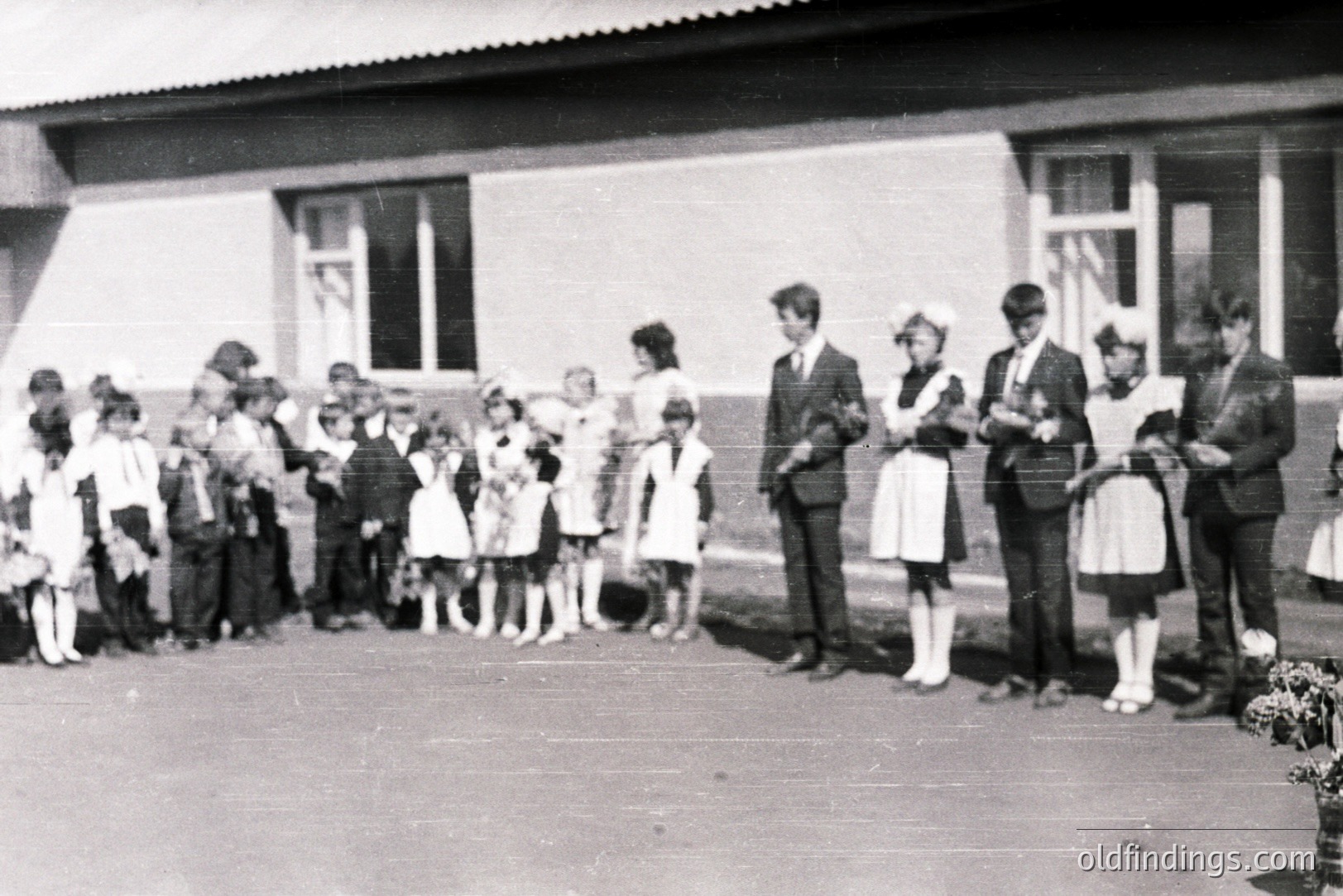 Group portrait outside a blocky, modernist building. A large group of children, some in uniform, stand alongside three adults, two appearing to be nurses and one man in a suit. Likely a school or institutional gathering, possibly Bulgaria, 1970s. A commemorative flower arrangement rests on the ground.