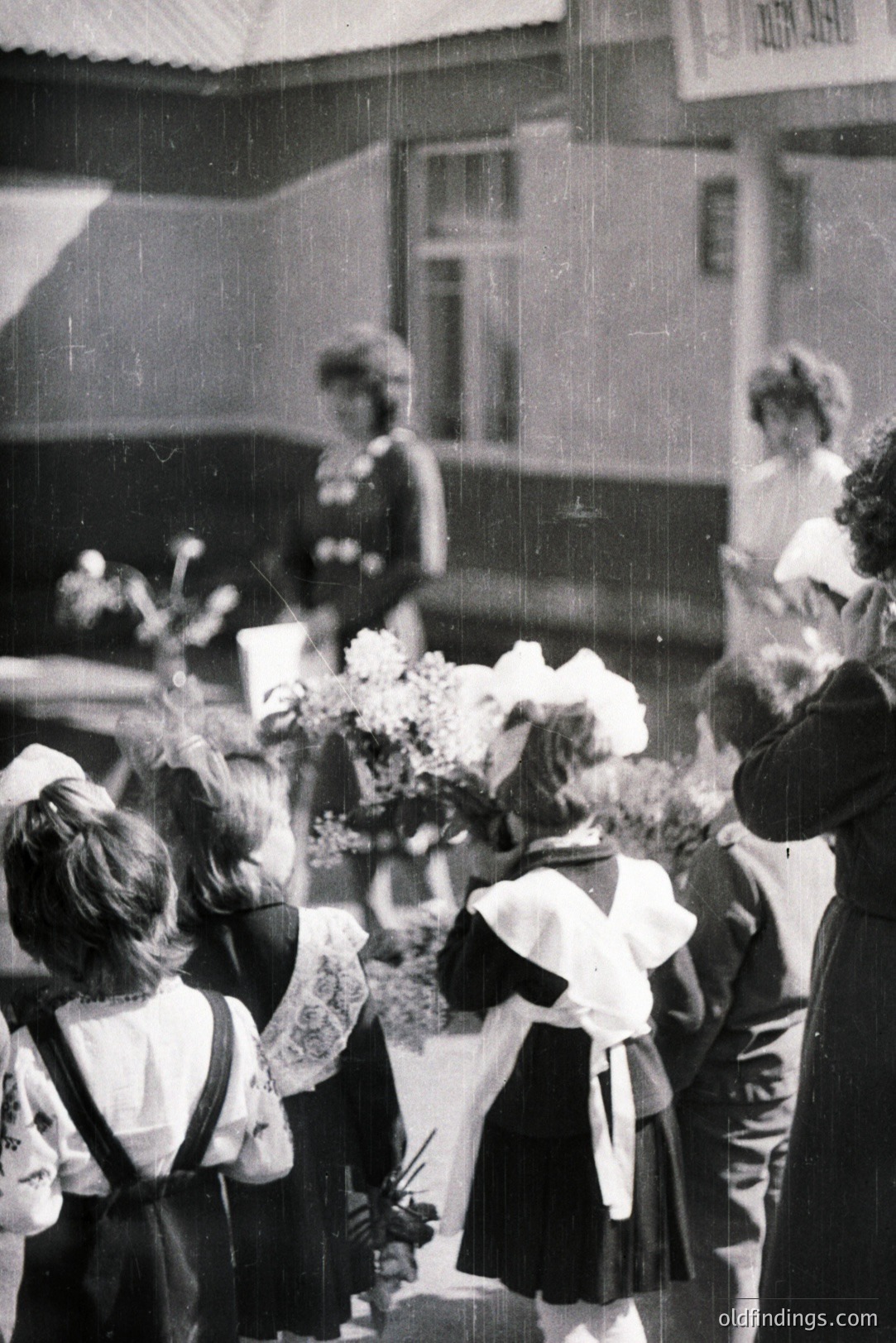 Group of children, many in traditional or folk attire, present bouquets to adults near a building. Appears to be a school or community event. Possible Eastern European setting, circa 1960s-1970s based on clothing and photo quality. Strong archival value for cultural studies.