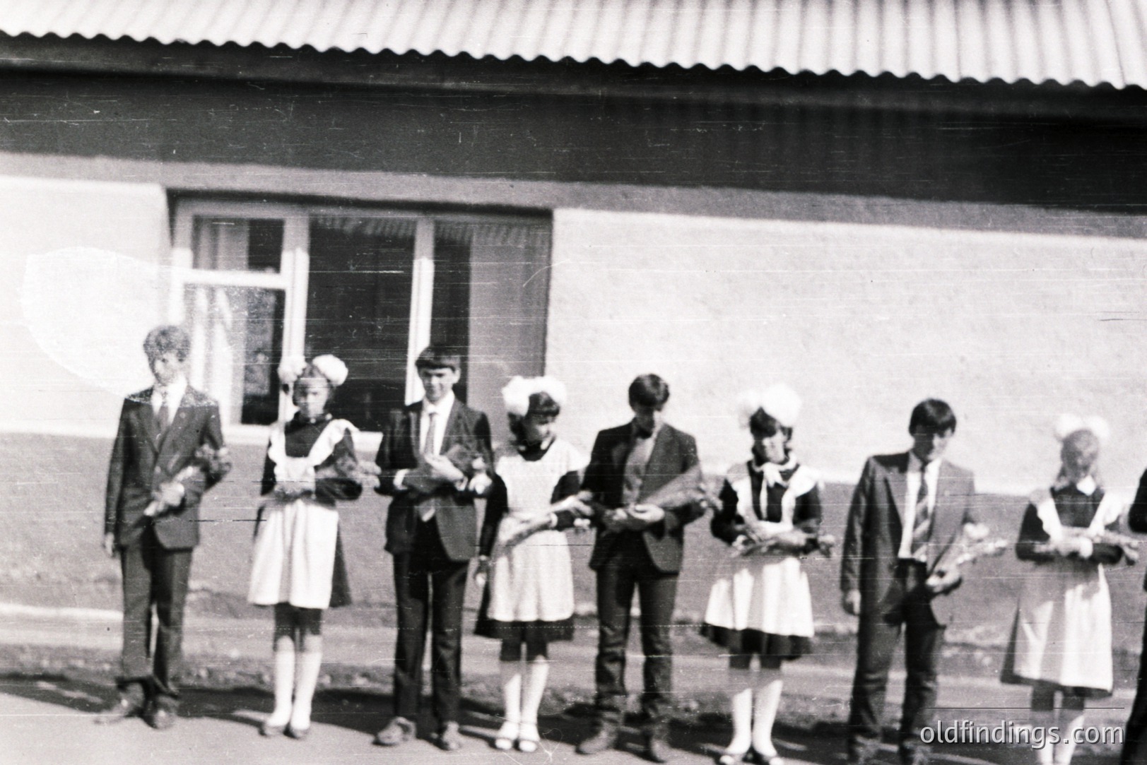 Six people pose in front of a building with a visible roof overhang and a single window. Four individuals wear vintage-style, short-sleeved dresses and sailor-like hats; two are dressed in suits. Likely a school or cultural event, captured circa 1960s-1970s. A commemorative photograph with potential historical value.