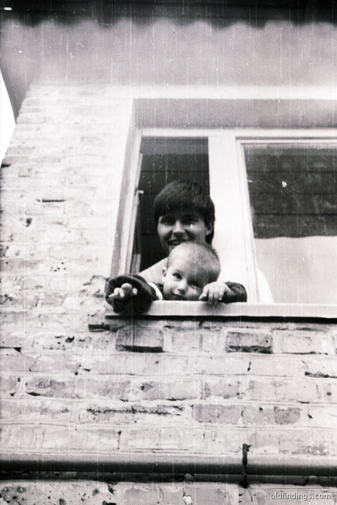 A young woman and baby peer out from a window with a brick facade. The woman has short hair and a cheerful expression; the baby looks directly at the camera. Likely a candid, informal portrait, evoking a sense of domesticity. Appears to be a mid-century snapshot.
