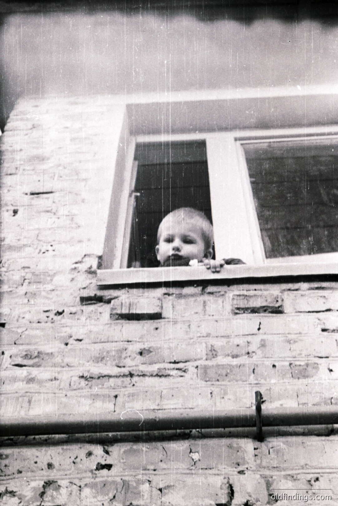 A young child peers from a window, framed by a brick wall and wrought-iron detail. Appears to be a candid moment, possibly a residential setting. Likely 1960s or 1970s based on photographic style and architectural details. Notable for its quiet, intimate feel.