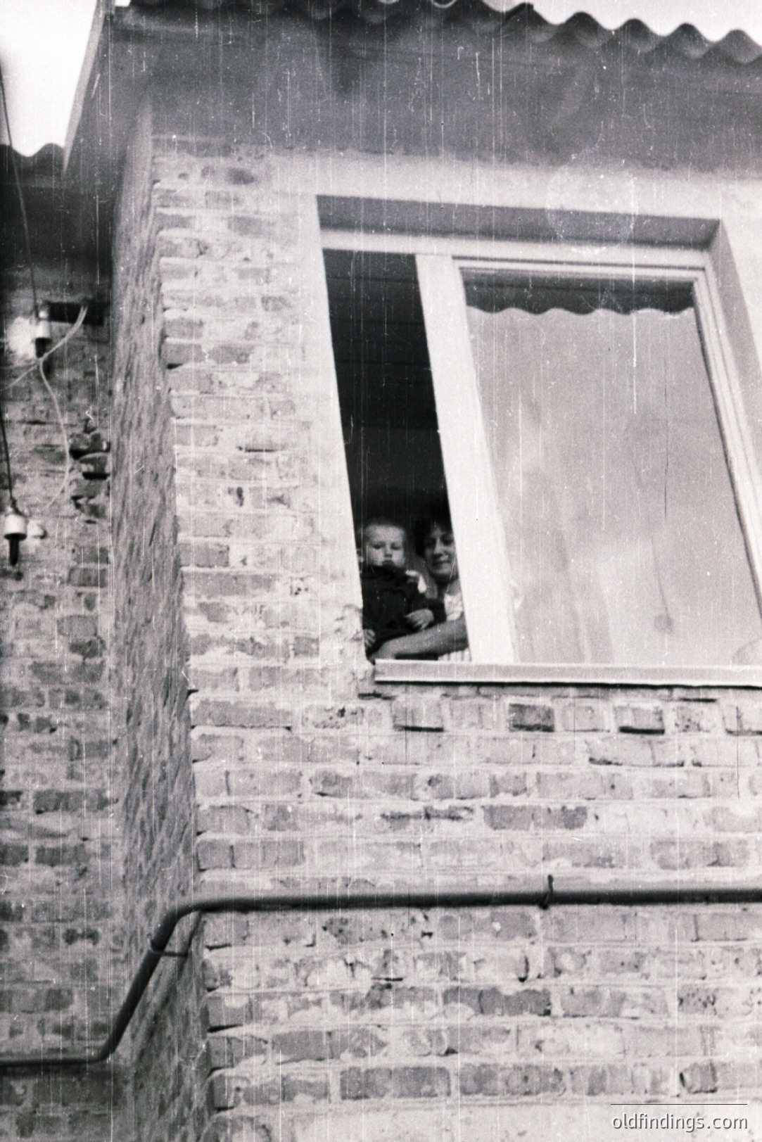 A young mother holding an infant sits posed in a window of a brick building. The exterior wall is visible, revealing aged brickwork and piping. Likely a domestic scene, hinting at family life. Appears to be a mid-century photograph, possibly 1950s or 60s. Good for historical research or design reference.