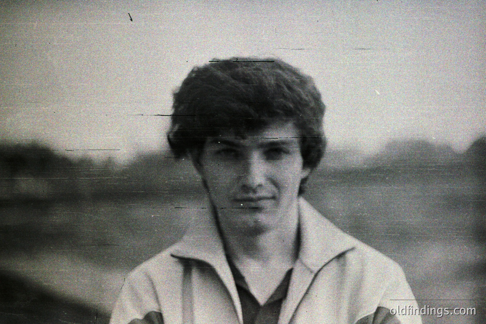 Formal portrait of a young man with dark, textured hair and a layered white shirt over a dark top. Natural, slightly blurred background suggests outdoor setting. Likely 1970s style. Image exhibits age-related degradation, including vertical scratches. Possibly a family snapshot or candid-style portrait.