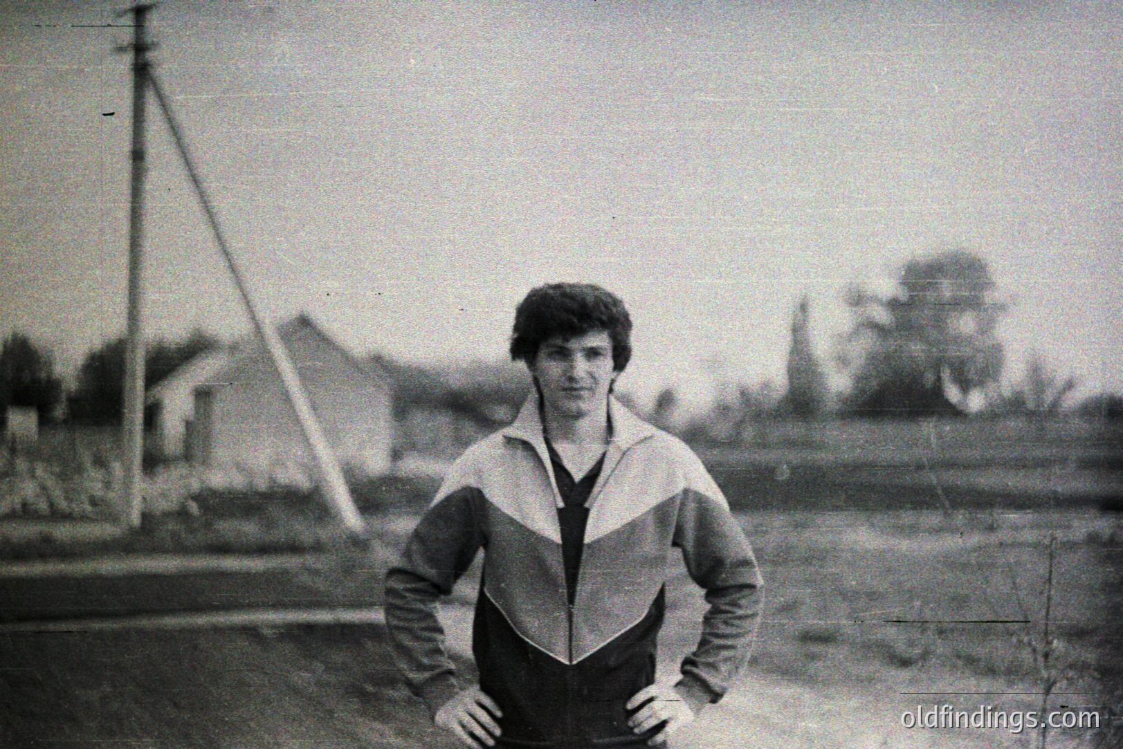 A young man stands with hands on hips, wearing a distinctive color-blocked tracksuit – black with white and red accents. Background shows a modest rural home and foliage, indicative of a casual, outdoor setting. Appears to be a snapshot from the 1970s. Potential for retro design reference.