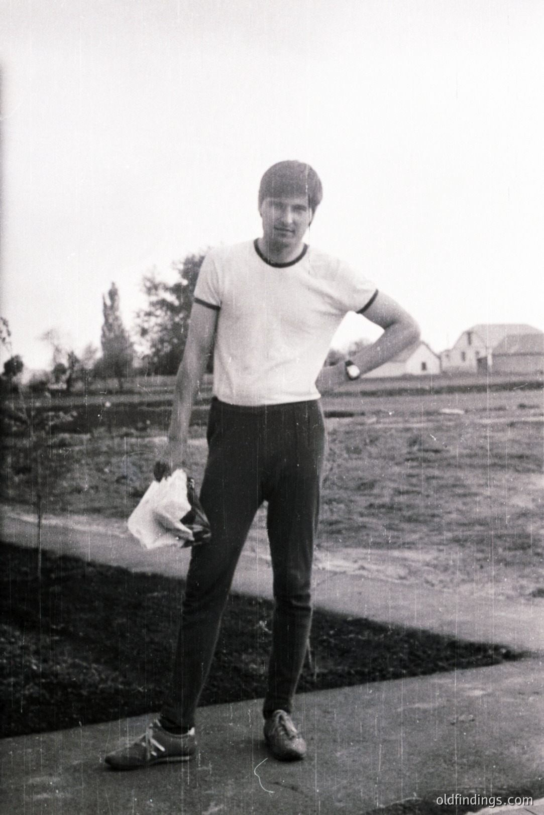 Young man stands outdoors, posed with one hand on hip, holding a folded cloth. He wears a white athletic tee, black trousers, and loafers. The location appears to be a rural area with a simple house in the background. Likely taken in the 1970s.