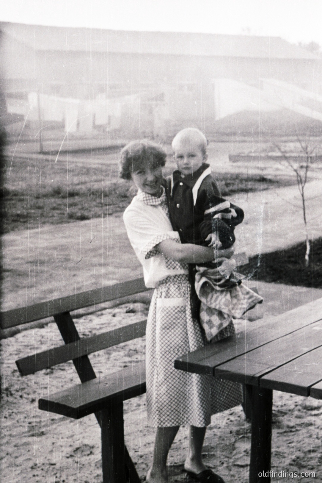 A woman in a patterned dress cradles a young boy on her hip, posed near a picnic table. Simple architecture and what appears to be a gravel yard backdrop. Likely a family portrait, possibly from the 1940s or 50s. Offers nostalgic charm and possible stock photography value.
