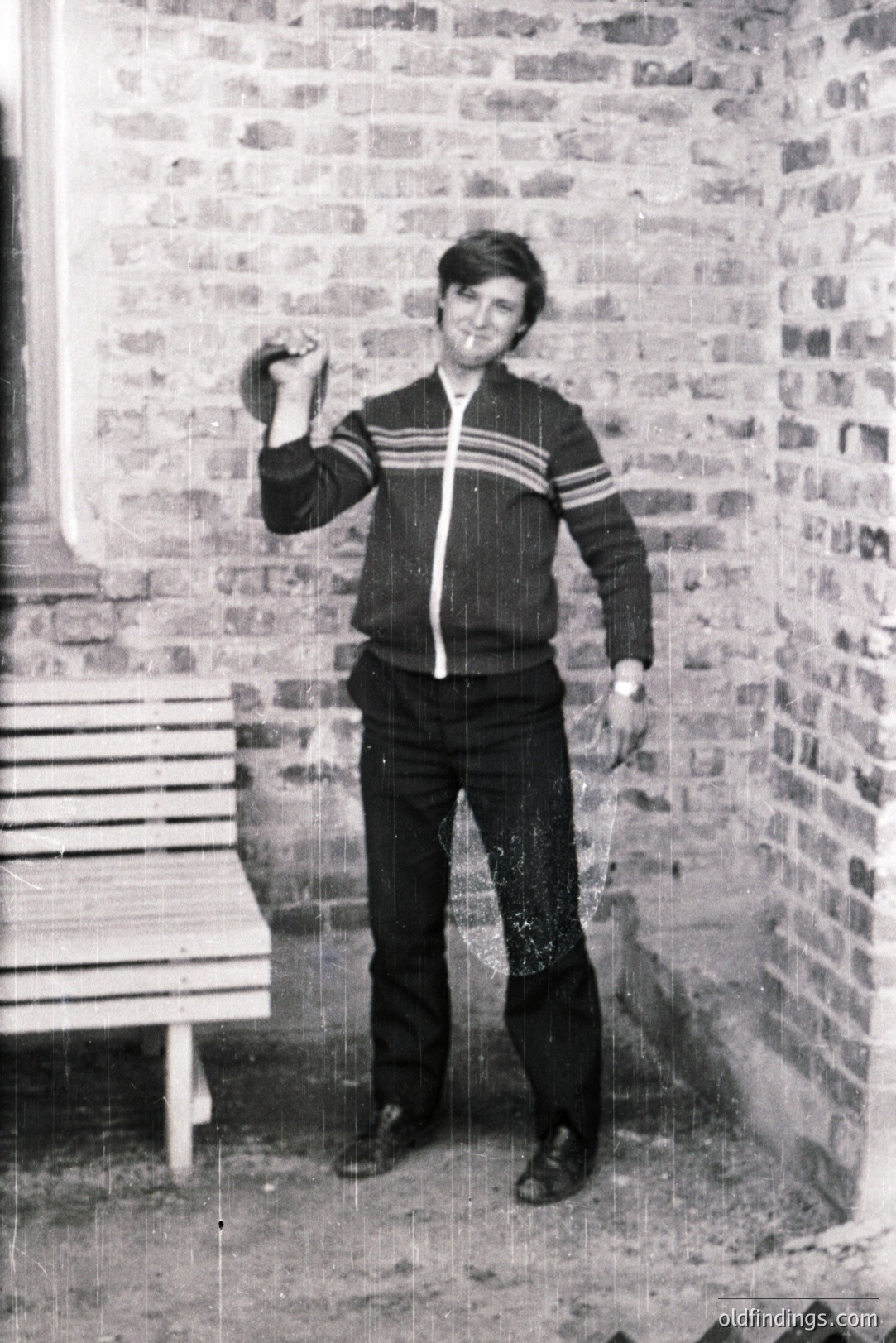 Man stands in an interior courtyard, holding a dumbbell. He wears a vintage tracksuit, bell-bottom trousers, and a watch. Brick walls and a bench are visible. Likely a fitness or lifestyle photo from the 1970s. Offers design/retro reference value.