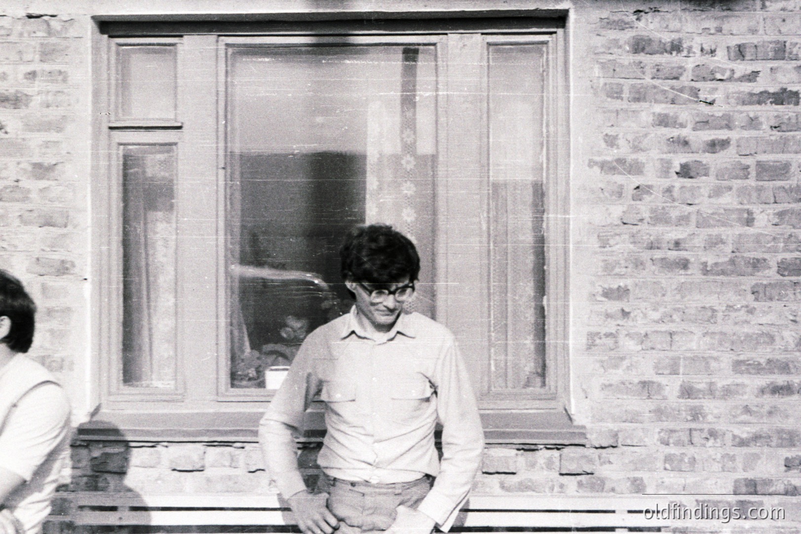 A man with dark hair and glasses stands outside a window in a brick building. He wears a collared shirt and trousers. The architectural detail suggests a mid-century modern style. Likely captured in the 1970s. A partial view of a person is visible on the left side.