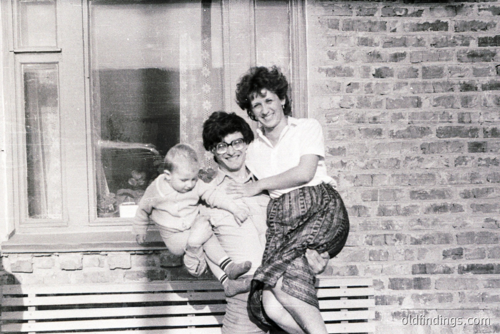 A smiling woman carries a baby outdoors, posed in front of a windowed brick building. The woman wears a short-sleeved shirt & patterned skirt; the infant is bundled in a cloth. Likely a candid snapshot, c. 1970s. Offers nostalgic family portrait potential.
