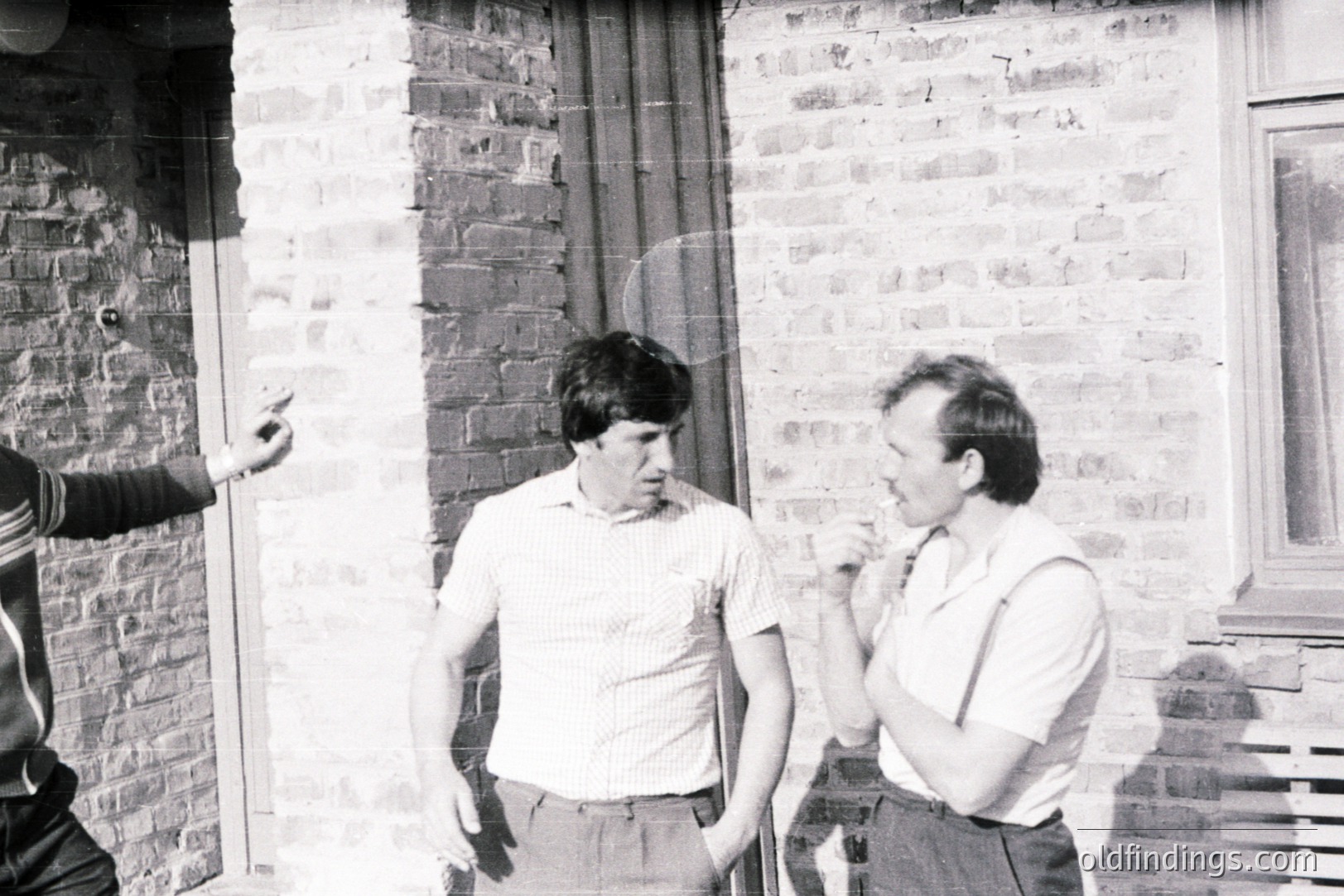 Three men stand outdoors near a brick wall, engaged in what appears to be a casual conversation. Two men are clearly visible – one with dark hair, the other with shorter, lighter hair, smoking a cigarette. Clothing style suggests the 1970s. Likely a candid snapshot.