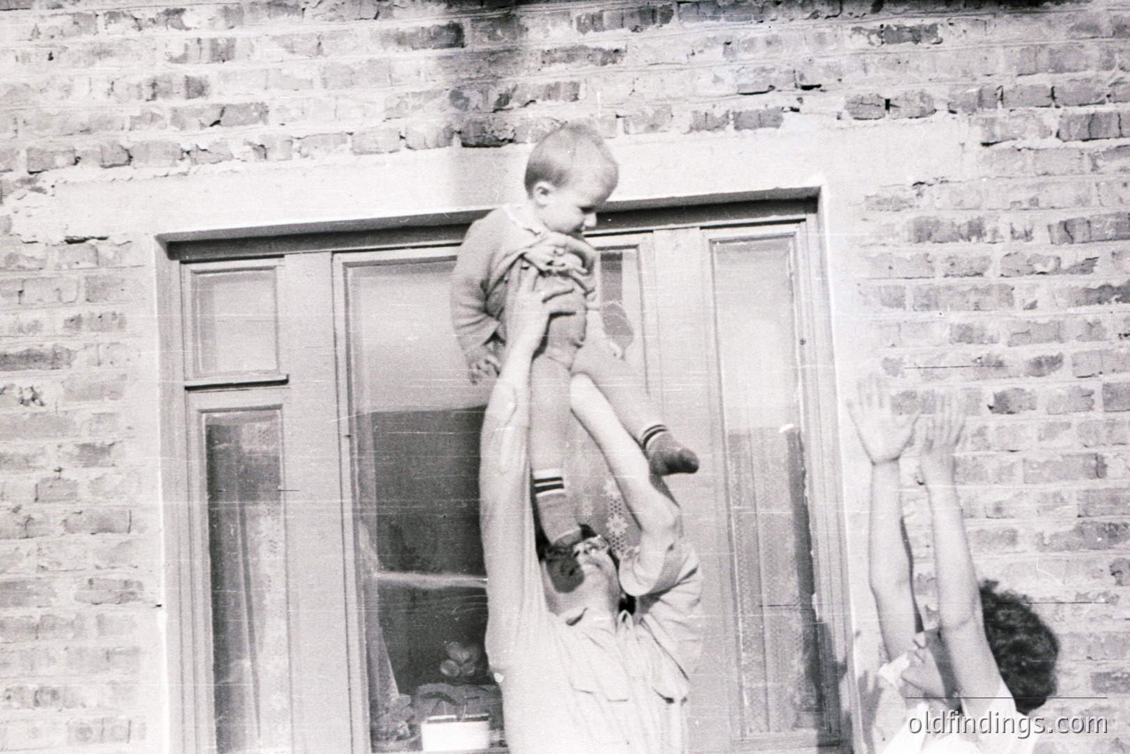 A child is held aloft by an adult in front of a brick building with a multi-paned window. The image’s texture suggests a vintage snapshot, likely from the 1950s or 60s. The child wears a knitted sweater. A casual, intimate moment captured in everyday domesticity.