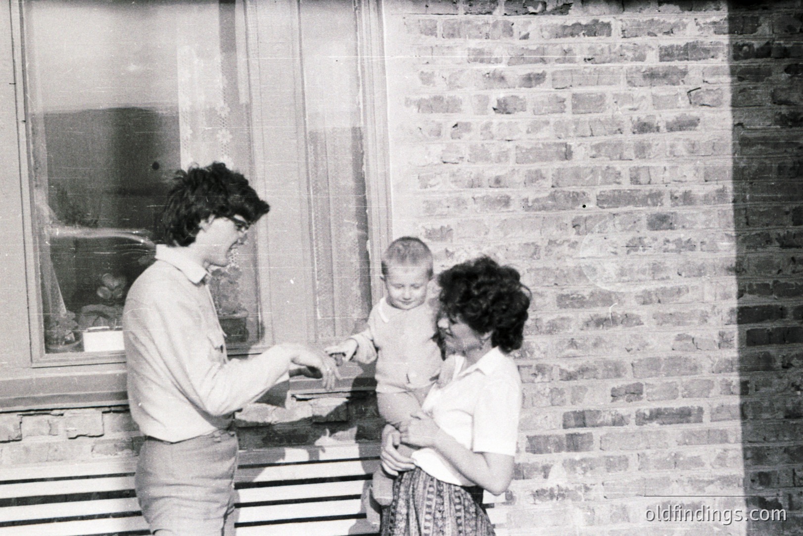 A candid family portrait showing a young boy being handed a treat by a man in bell-bottoms, while a woman holds him close near a brick wall and window. Likely a 1970s snapshot capturing a moment of connection & everyday life. Simple domesticity.