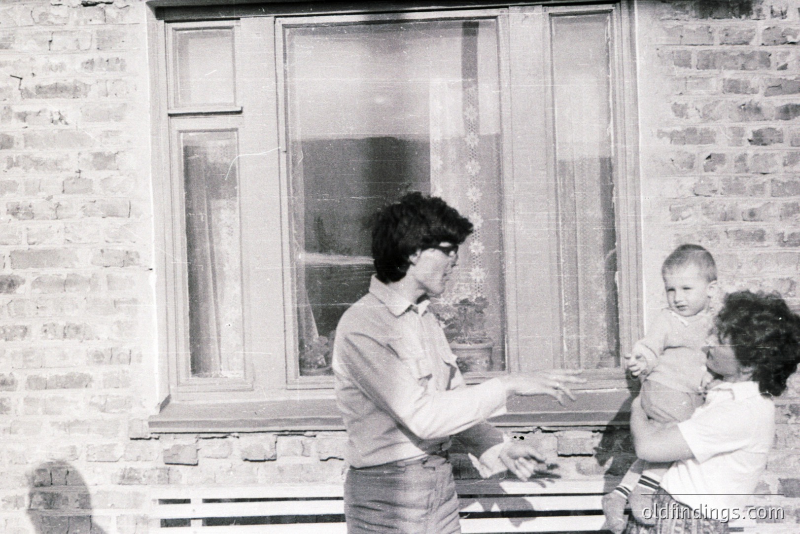 A candid black and white photograph shows a young boy being held by a woman outside a brick building. The woman and child are directly in front of a window with patterned curtains. The man next to the woman appears to be speaking with her. Likely a domestic scene from the 1970s.