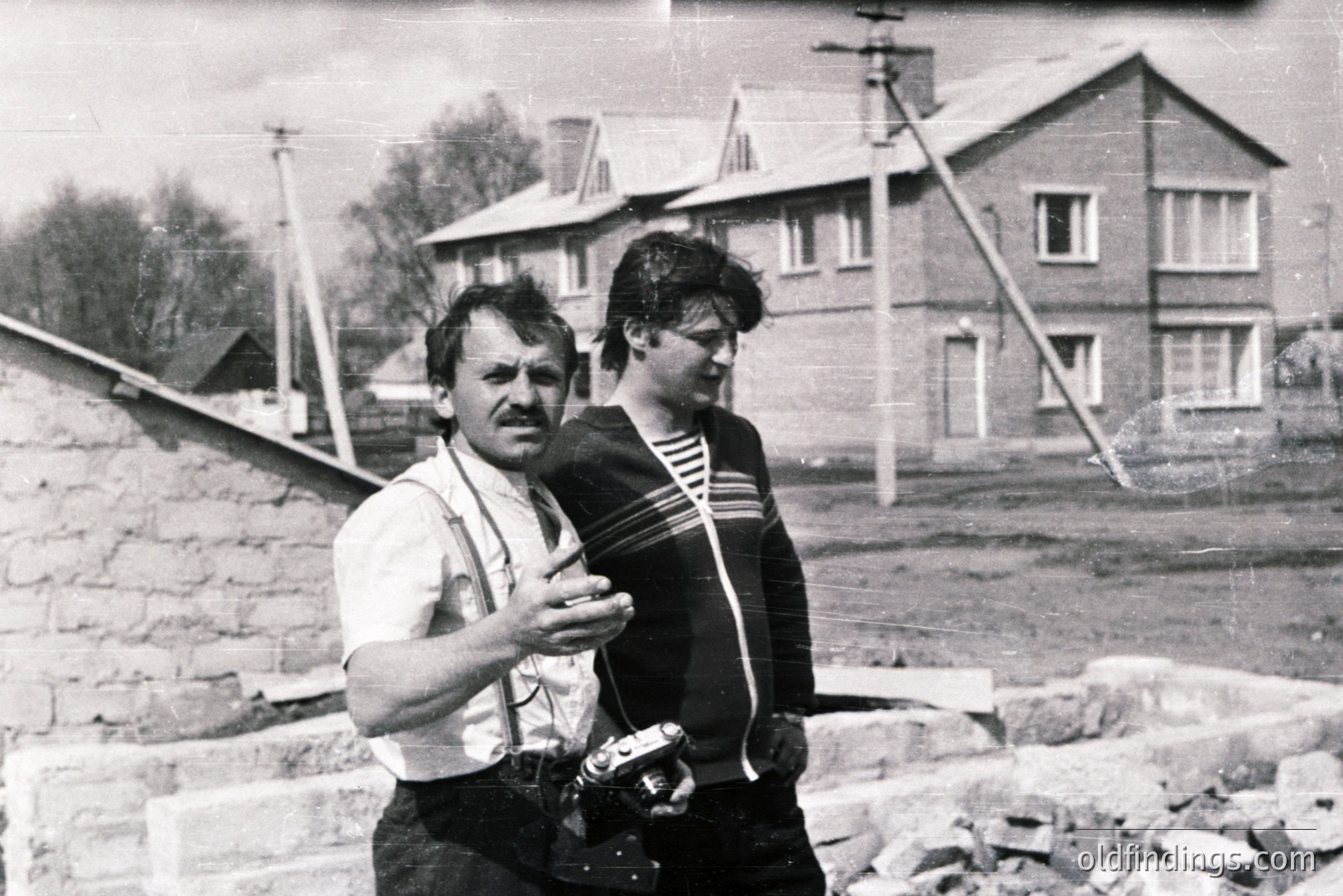 A man in workwear, suspenders, & a bricklayer, converses with a younger person in a striped sweater during what appears to be construction. A partially built brick wall & a row of houses are visible. Likely a candid, mid-century snapshot. Architectural documentation or family archive potential.