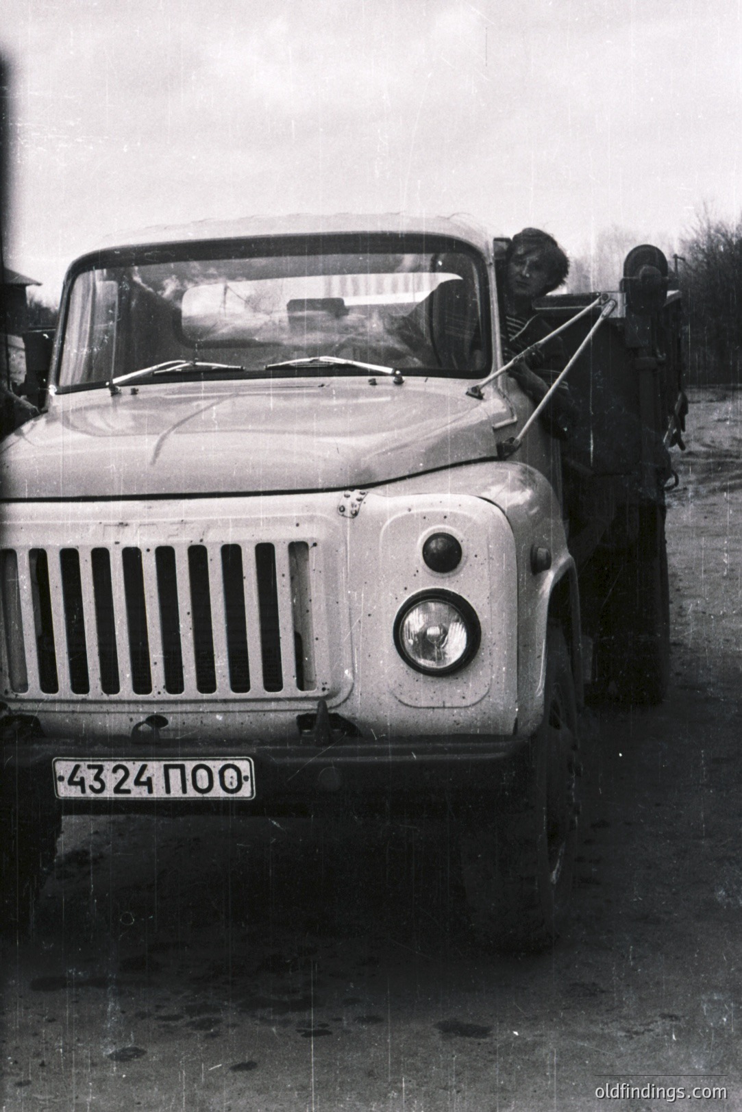 A utility truck, likely a Soviet-era UAZ-469, drives on a wet, gravel road. A man hangs from the door, smiling, while the driver is visible. License plate reads "4324 ПО0." The vehicle’s robust design indicates use on rough terrain. Photograph captures a moment of casual transport.