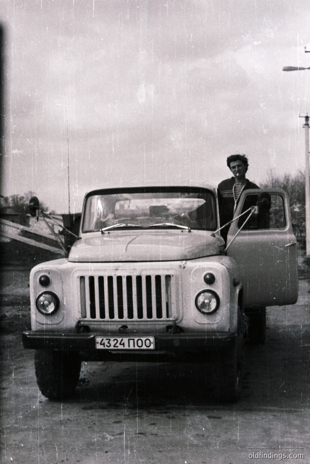 A young man stands in an early model UAZ-69 jeep, door ajar, appearing to smile at the camera. The vehicle's Cyrillic license plate is visible. Likely Soviet-era, captured in a stark black and white image, possibly a personal snapshot. Represents a period of utilitarian vehicle design.