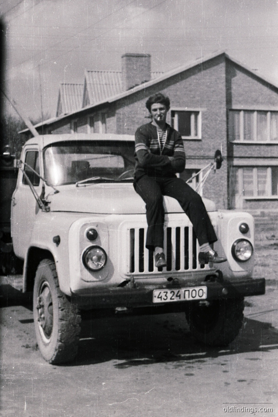 A young man in a striped shirt sits atop a vintage Soviet-era truck, likely a UAZ-452. The vehicle's license plate reads "4324 ПОО." The scene is framed by a modest, brick-clad house. Likely Bulgaria, 1970s. Represents everyday life under socialist influence.