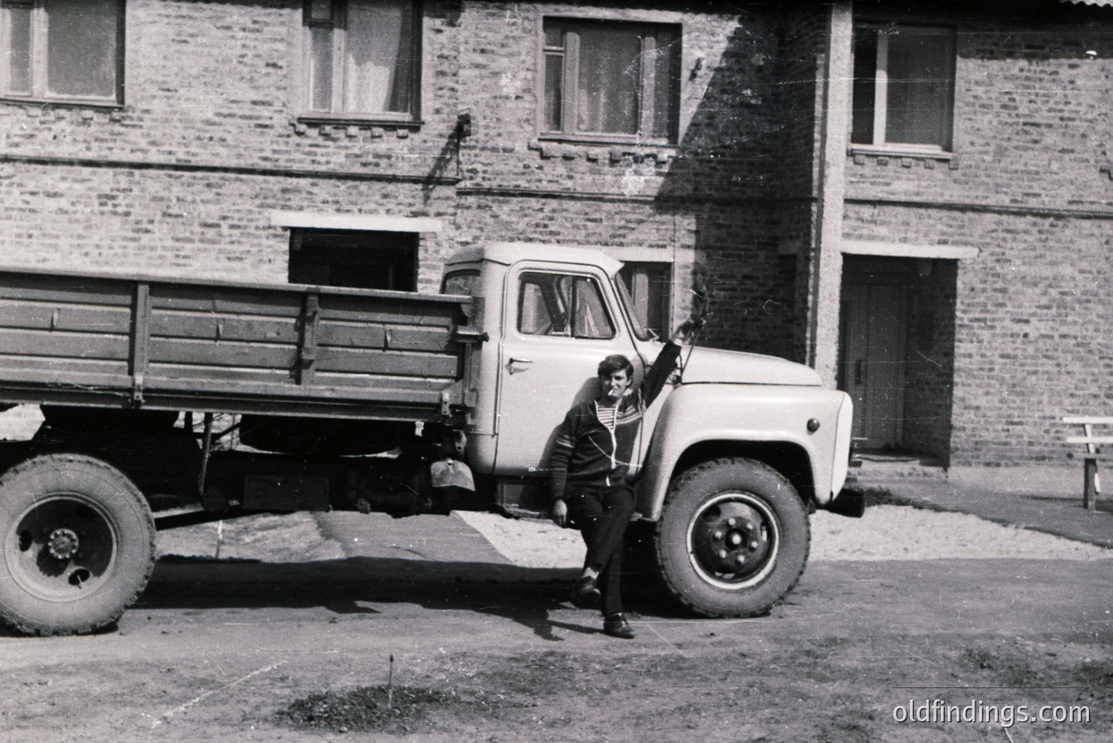 A young man stands leaning against a utilitarian Soviet-era truck, likely a UAZ-452, in front of a brick apartment building. The building’s architecture suggests a mid-20th century Eastern European design. The image exhibits grainy quality typical of film photography. The man’s clothing and the truck’s design point to a 1960s-1970s timeframe.