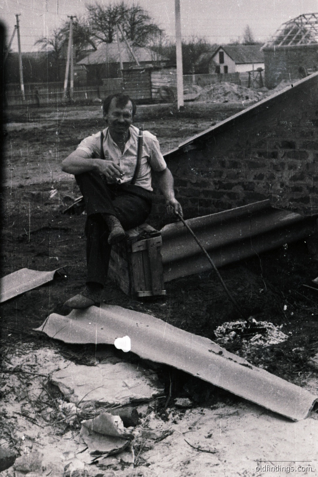 A construction worker, likely Bulgarian, sits on a wooden crate amidst debris and roofing materials. He wears a short-sleeved shirt, suspenders, and work boots, holding a tool. Stone wall & unfinished structure in background. Appears to be the 1960s/70s.