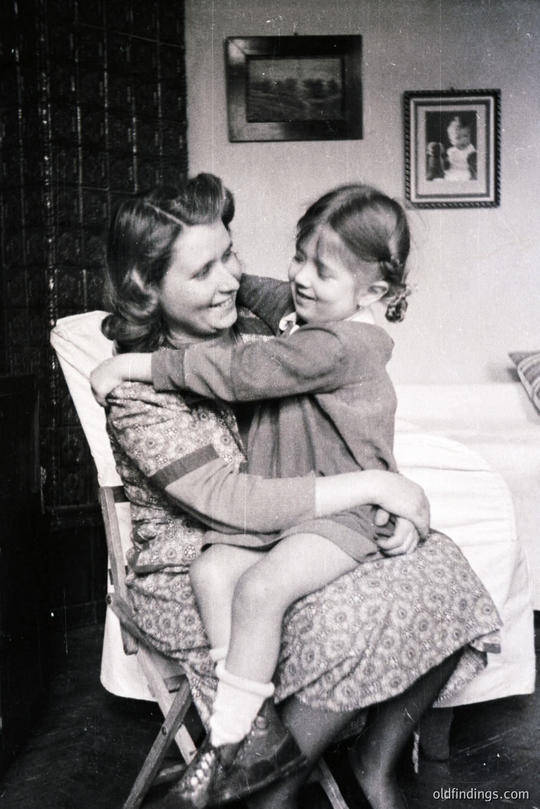 A mother and daughter share a loving embrace on a folding chair. The woman, dressed in a patterned dress, smiles warmly at her daughter, who wraps her arms around her. The setting appears to be a home interior, indicated by the framed artwork on the wall and wallpaper. Likely mid-20th century. A charming, tender moment captured in time.
