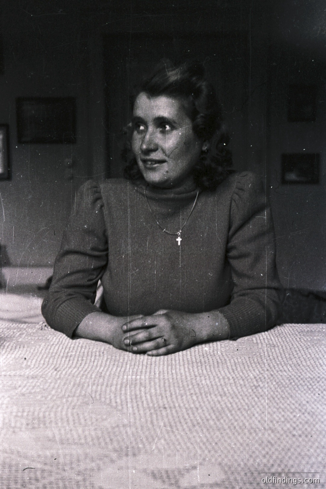 Vintage portrait of a woman seated on a bed, hands clasped. She wears a dark turtleneck sweater, a delicate necklace with a cross. The backdrop reveals a simple, possibly rural interior with framed pictures. Likely 1960s-70s. A study in everyday domestic life.