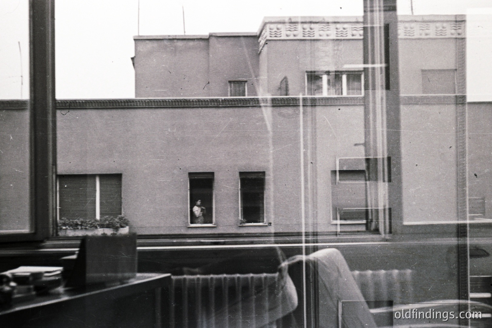A monochrome image shows an exterior view through a window, likely an office or apartment. Visible is a modernist building with a flat roof and symmetrically placed windows. A figure stands in one window. Includes visible window frame and interior elements. Likely 1960s-1970s.