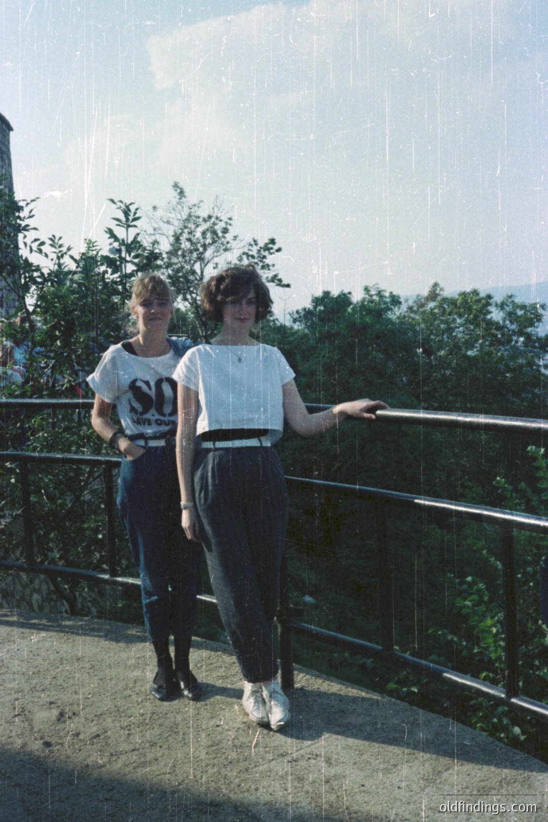 Two young women, dressed in 1980s streetwear, stand on a weathered concrete overlook. One wears a graphic tee, the other a cropped white shirt & high-waisted jeans. A lush, green landscape is visible behind the railing. Distinctive film grain and slight discoloration indicate age. Likely a casual snapshot.