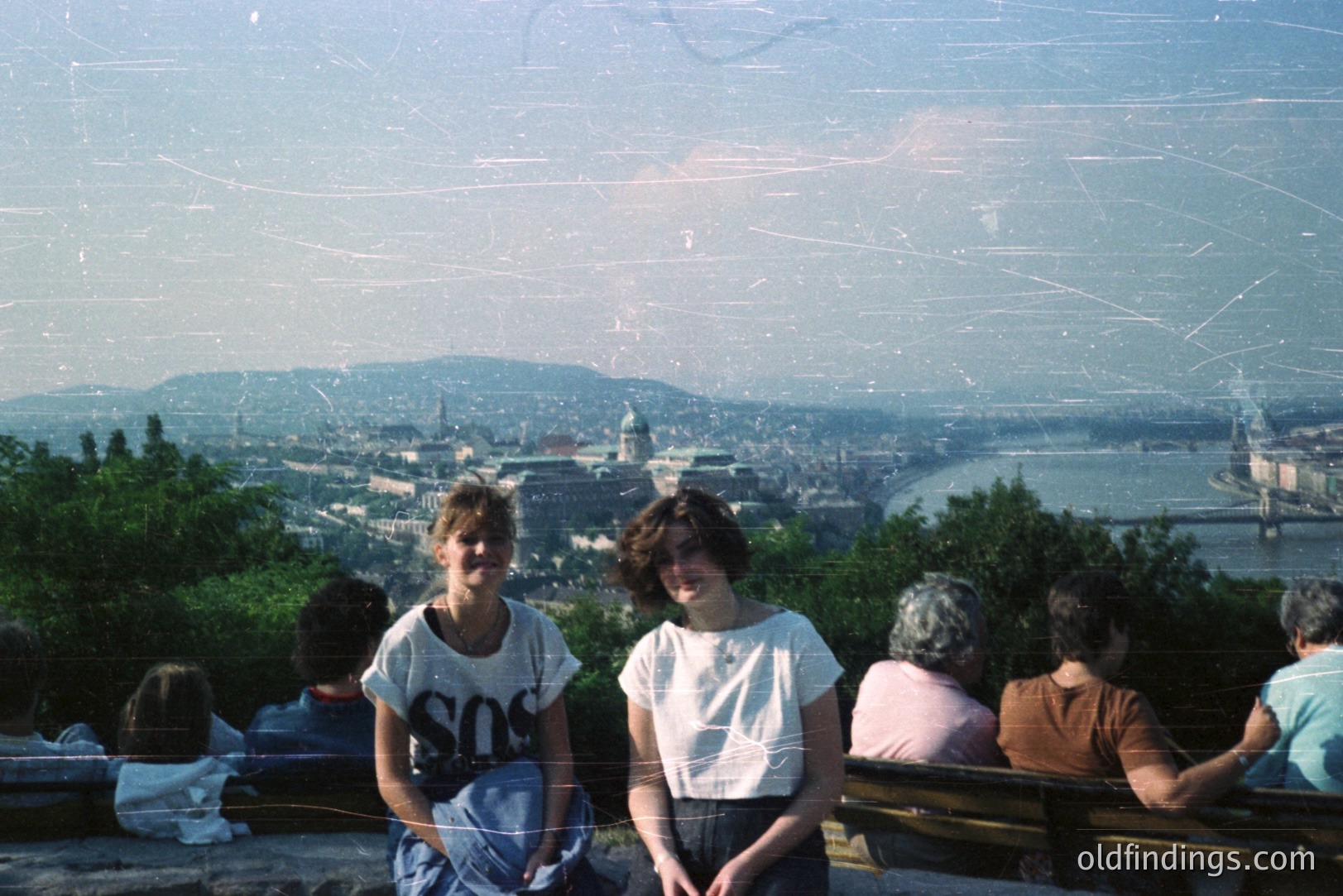 Two young people sit on tiered stone benches overlooking a cityscape, likely Budapest, Hungary. The panoramic view includes the Danube river and architectural details – domes and rooftops. Image exhibits characteristics of 1980s film photography: grain, color saturation, and visible scratches. A group of people sits behind the subjects.