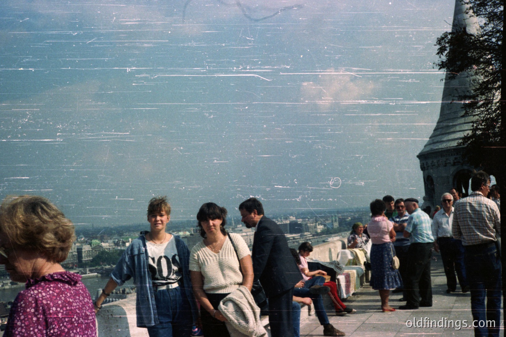 Crowded viewing platform, likely Paris, France. People observe a distant cityscape; the Eiffel Tower dominates the right frame. Fashion suggests the 1980s – denim jackets, high-waisted jeans, and oversized shirts. Noticeable film grain and surface scratches. Tourist scene, potential stock photo reference.