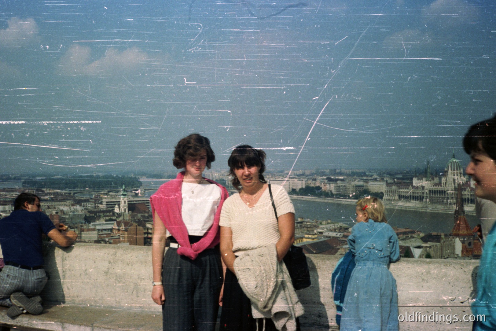 Two young women, likely tourists, pose with the Budapest skyline—Parliament building & Danube River—in the background. Photo appears to be from the 1970s/80s, judging by clothing & film grain. Vintage aesthetic, visible scratches add character.