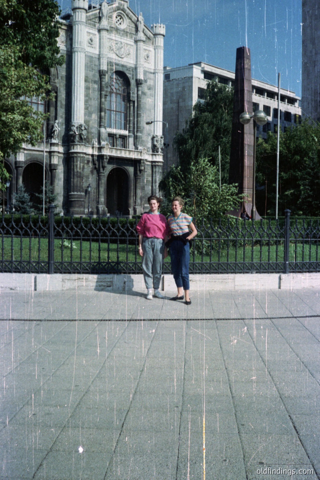 Two young people stand before a grand, ornate building exhibiting neoclassical architecture. The building features multiple arched windows, sculptures, and decorative detailing. A tall monument and a modern office building are visible in the background. The subjects’ attire suggests the 1980s. Likely taken in Mexico City.