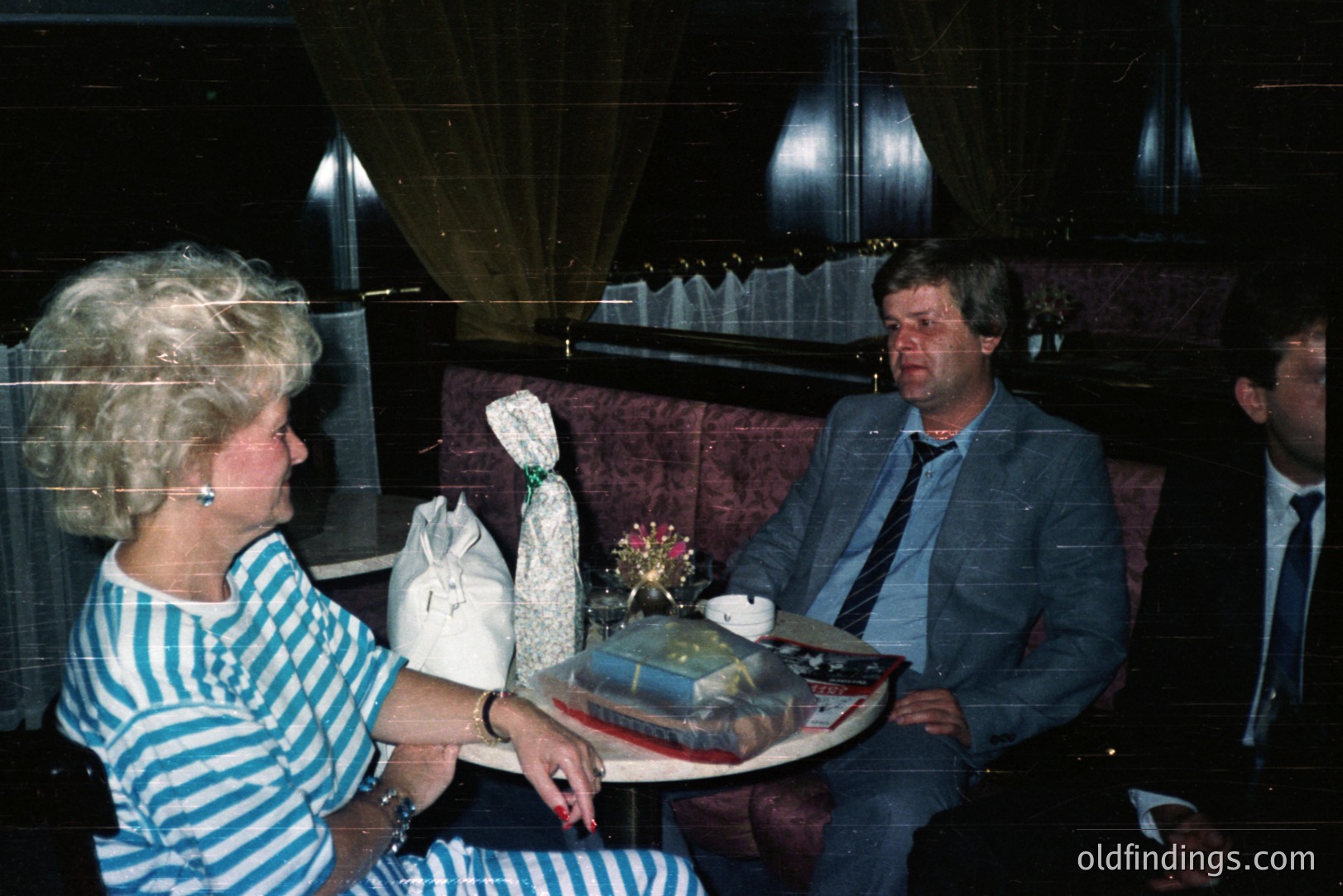 Seated individuals in a plush, dimly-lit interior, possibly a hotel lounge or formal dining area. A woman in a striped shirt, and a man in a gray suit are engaged in conversation. A third man sits partially visible. Decorative floral arrangement and boxed gifts are on a table.