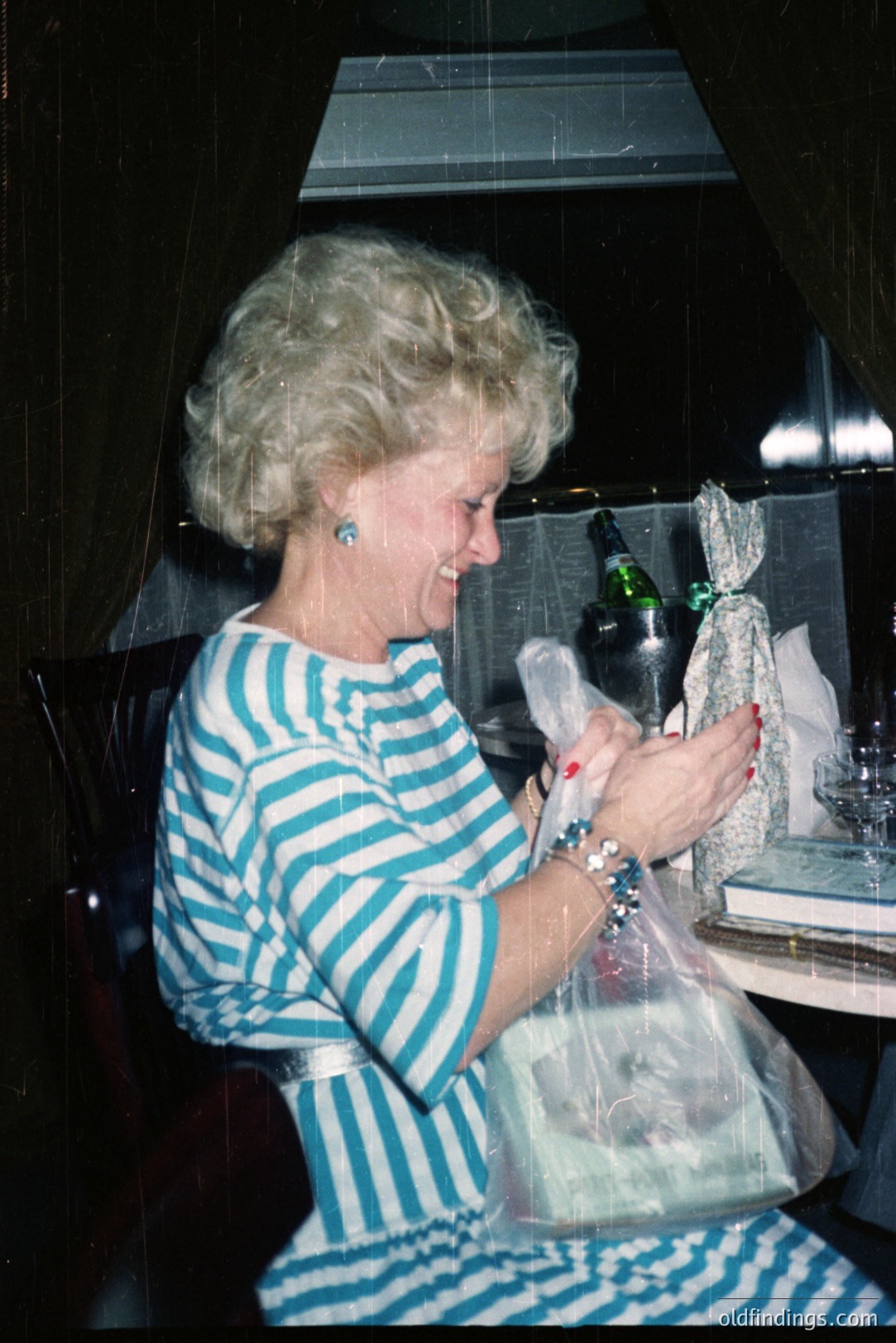 A smiling woman, sporting a voluminous, blonde hairstyle, appears delighted while receiving a wrapped gift. She wears a blue and white striped dress, silver bangles, and small earrings. A bottle of sparkling wine sits on a table with linens. Likely a festive occasion, late 1970s style.