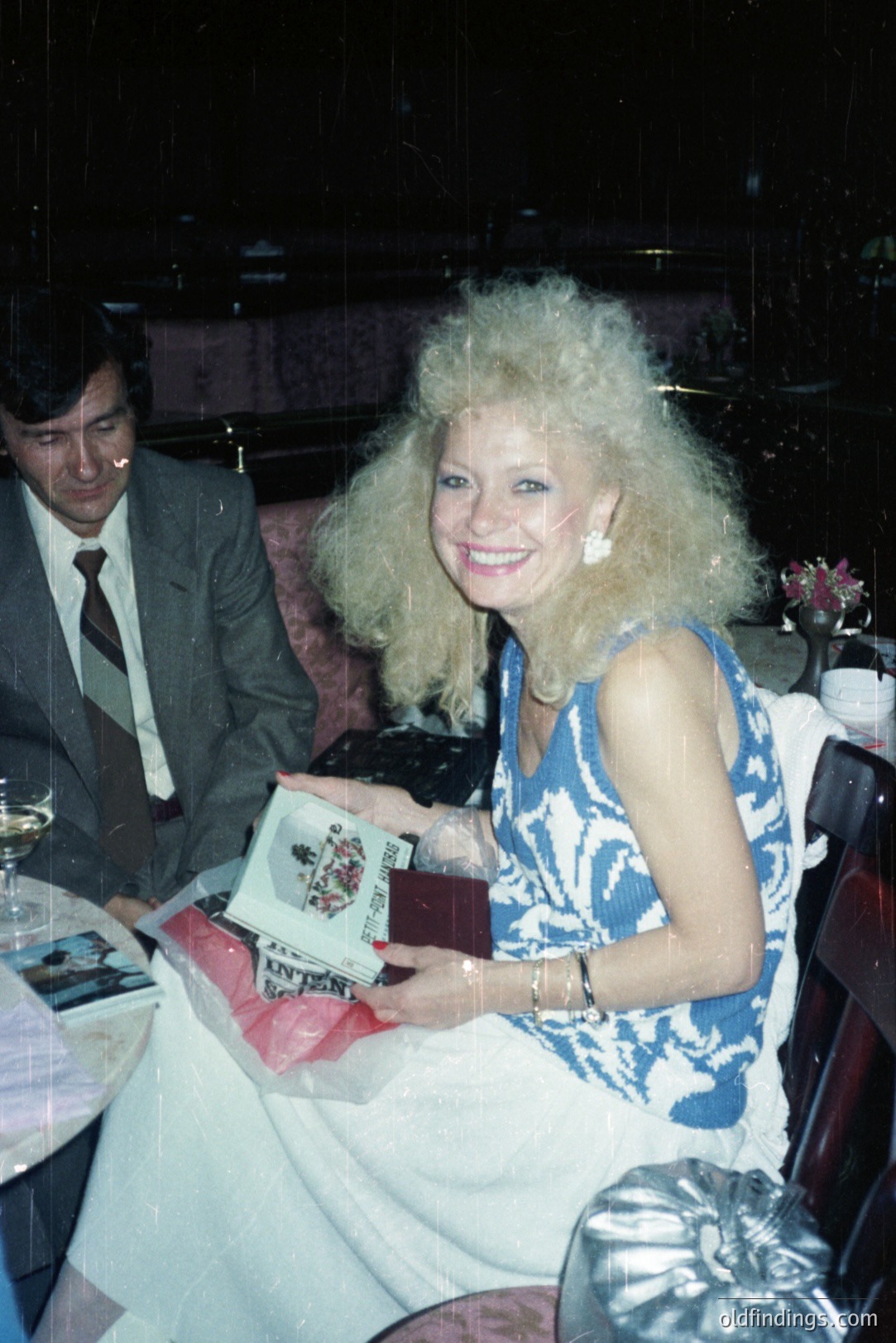 A smiling woman with voluminous 1980s hair holds a wrapped gift and a book, seated at a restaurant table. A man in a suit sits beside her. The decor features dark wood and floral arrangements. Likely from the 1980s. Commercial value: vintage style reference.