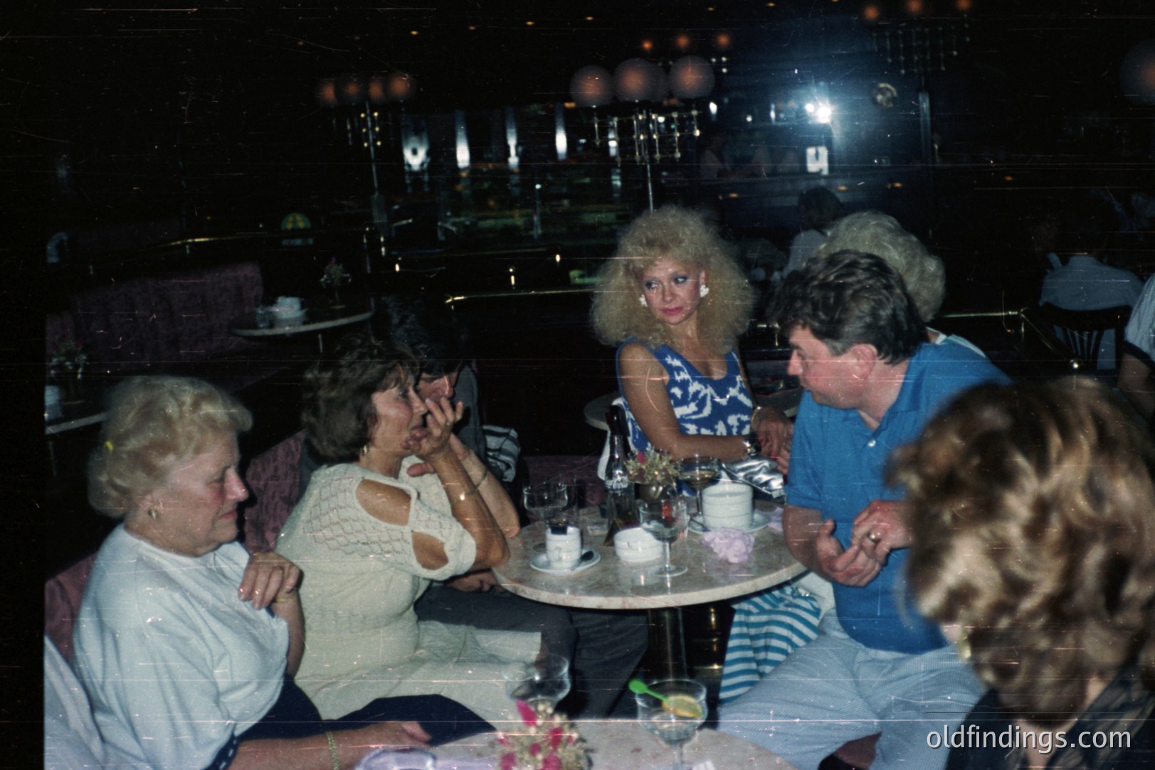 Group of people socializing at a table in a dimly lit, upscale restaurant or lounge. Visible are women with voluminous hairstyles and men with distinctive 1980s fashion. Likely a celebration or special occasion.