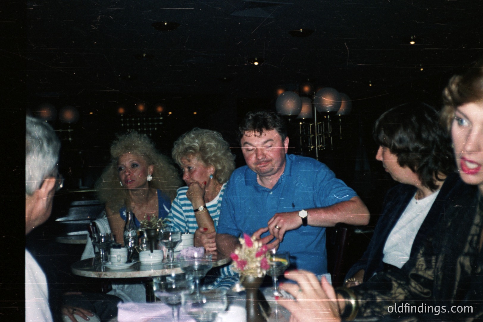 A group of people sit around a table laden with glassware, likely in a restaurant or bar. The composition indicates a candid moment – a flash photograph. Fashion suggests late 1980s or early 1990s. Visible elements include elaborate hairstyles, bold makeup, and typical casual attire. Potential stock photo value for retro aesthetics.