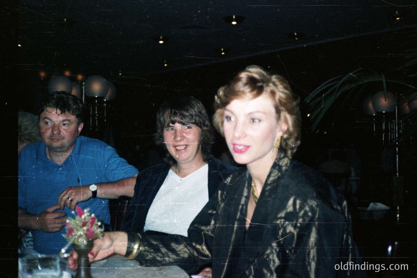 Three people stand in what appears to be a dimly lit, upscale event space. A man in a blue shirt stands to the left, two women are in the foreground. The woman on the right wears a patterned, dark jacket with a bold red lip. Likely 1980s, judging from the hairstyles and clothing. The event seems celebratory or formal.